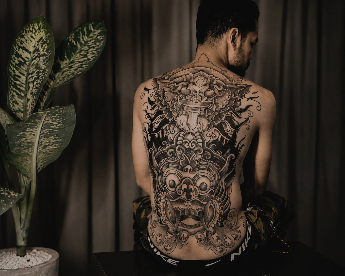 A man with a large, intricate black tattoo of a traditional dragon or demon face covering his back and upper body, sitting against a dark curtain background, with a large green leafy plant on his left.