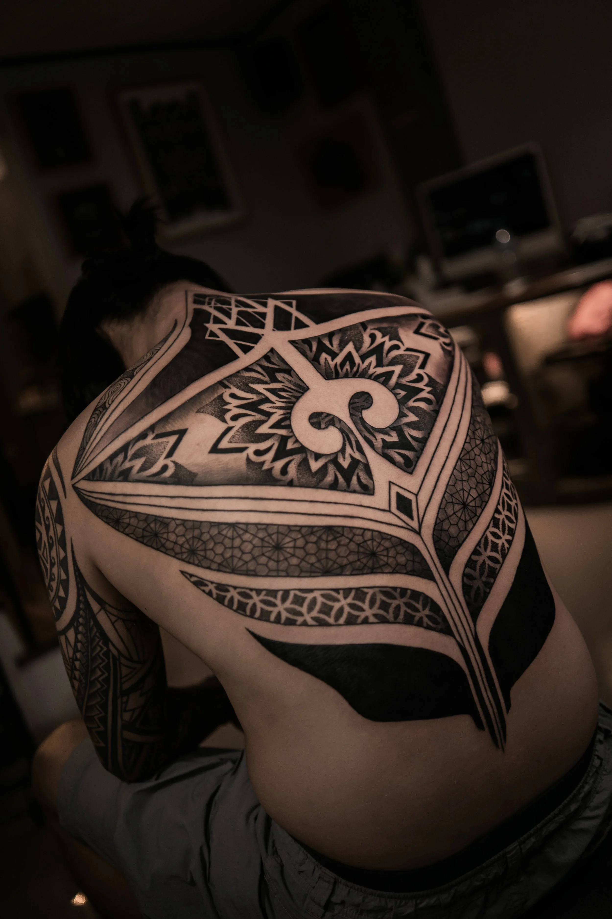 A person's upper back and shoulder covered in an intricate black and gray geometric tattoo with floral and symmetrical patterns.