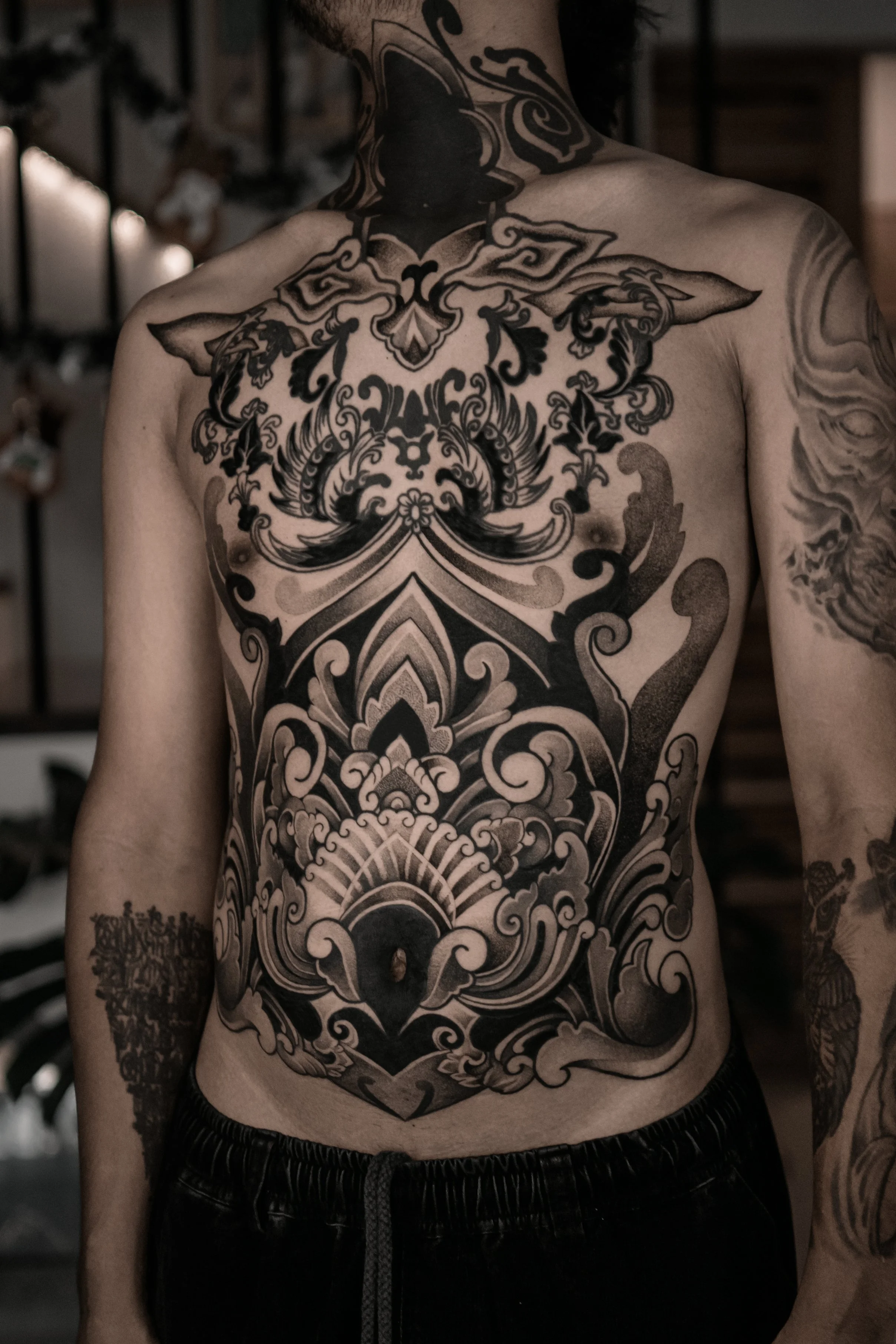 A person with extensive black tattoos on their chest, arms, and neck, featuring intricate patterns and designs.