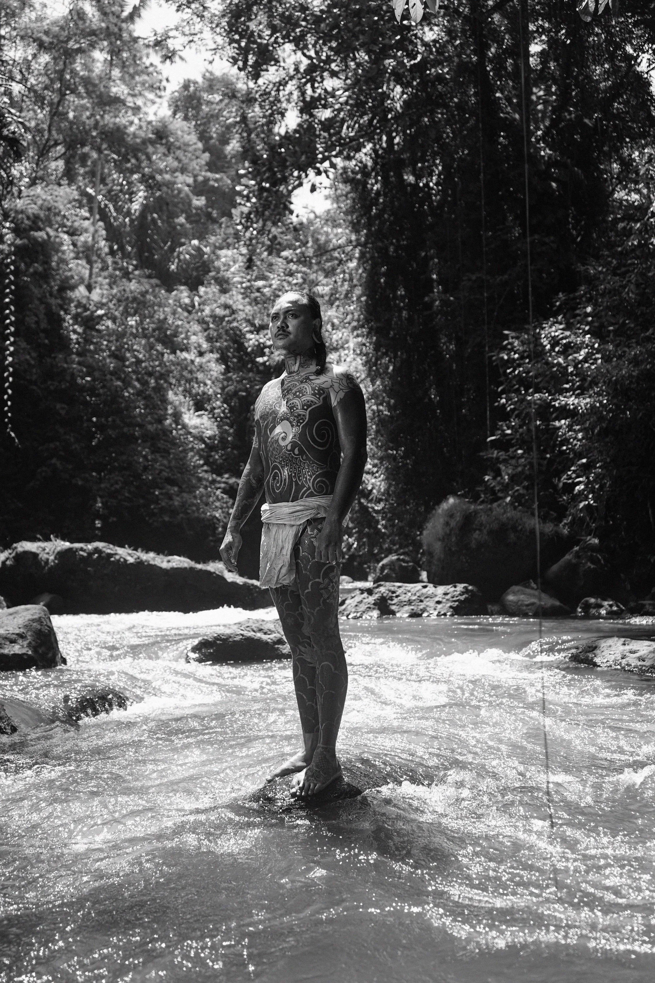 A person with intricate tattoo art on their body standing in a shallow river stream with rocks, surrounded by dense trees and foliage, captured in black and white.