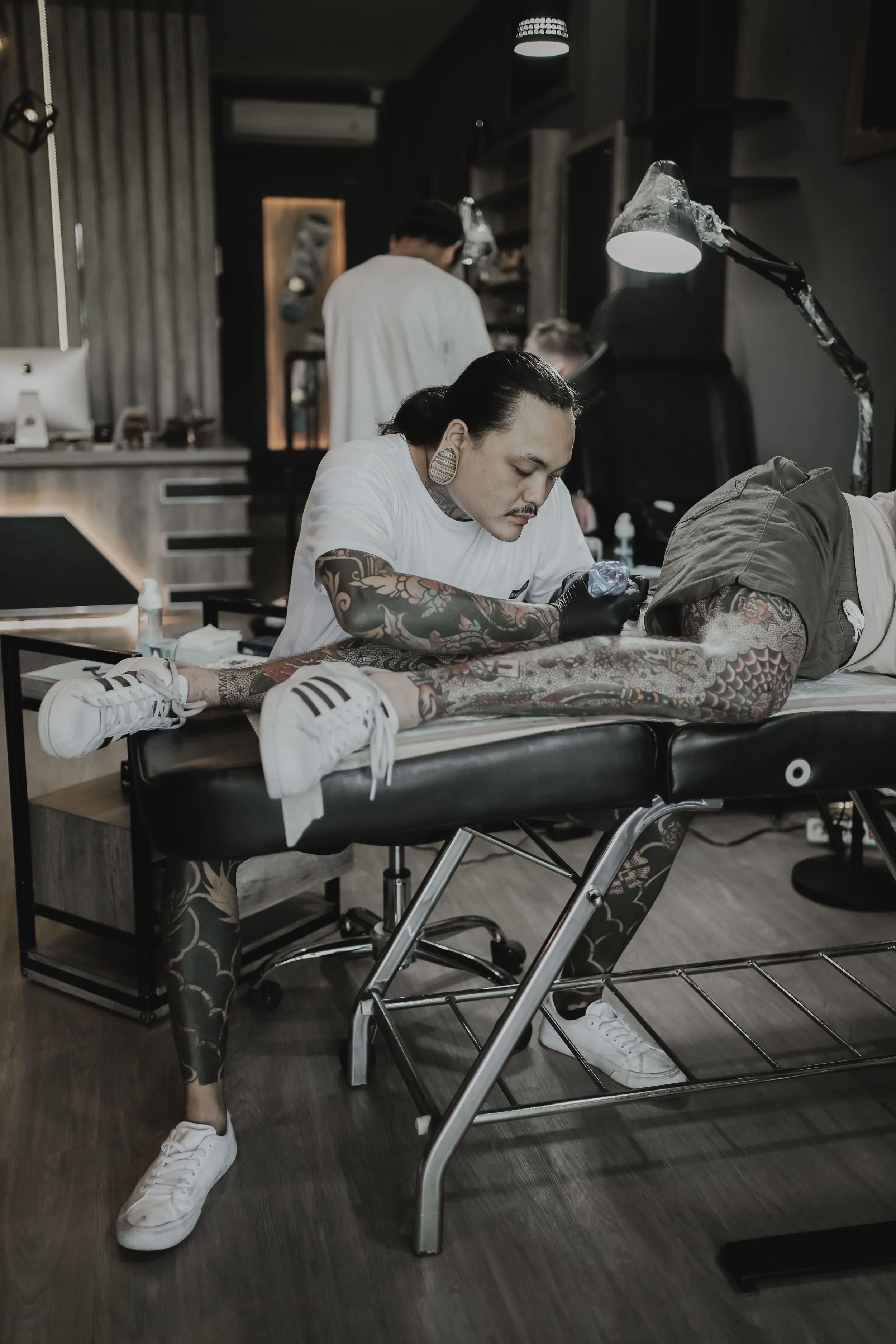 A tattoo artist working on a person's arm at a tattoo studio with a client lying on a table wearing white sneakers, tattoos visible on his legs.