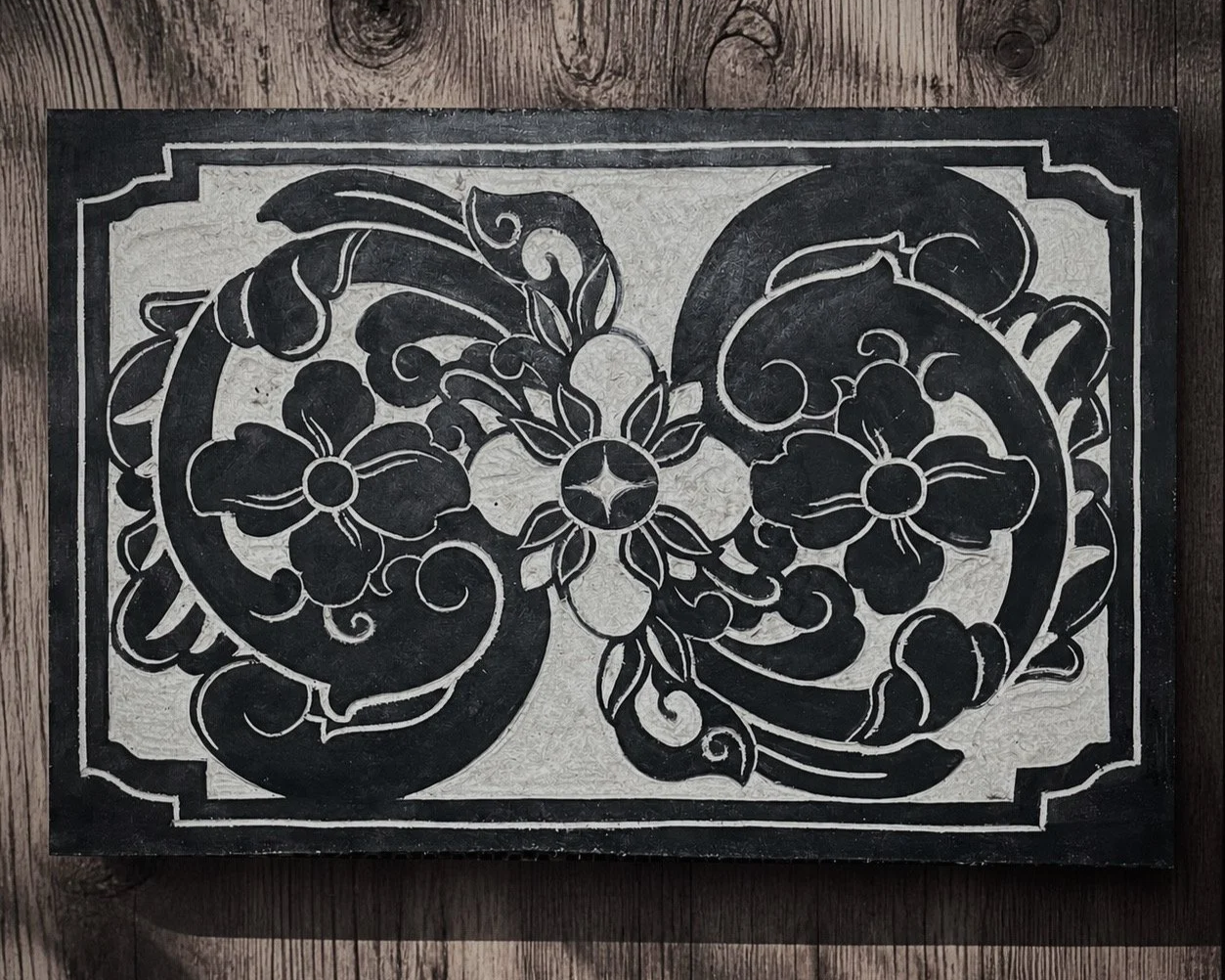 Black and white decorative tile with floral and scroll patterns on a wooden surface.