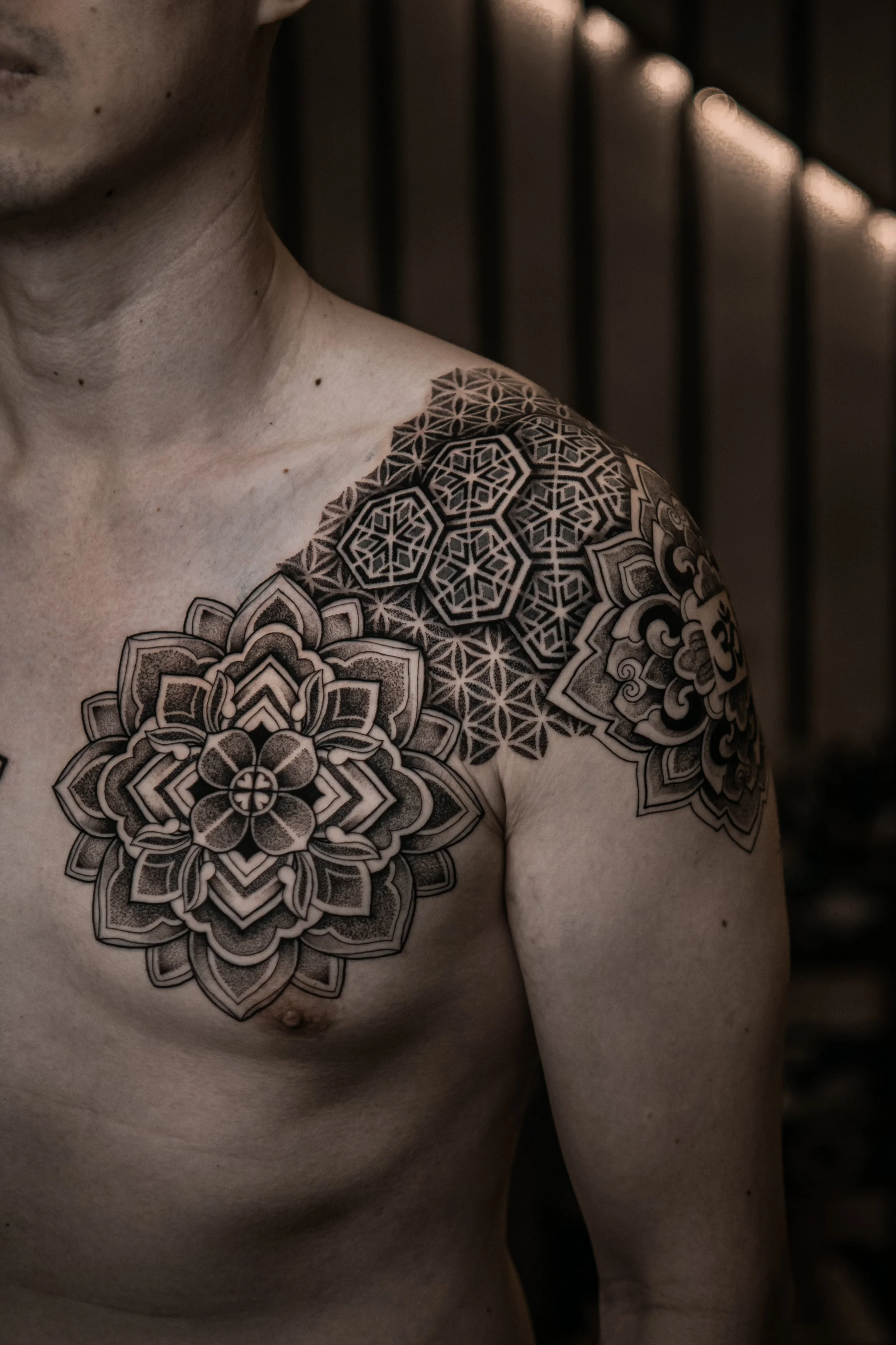 Close-up of a person's upper chest and shoulder showing intricate black geometric and floral tattoos on left shoulder and chest, with visible skin details.
