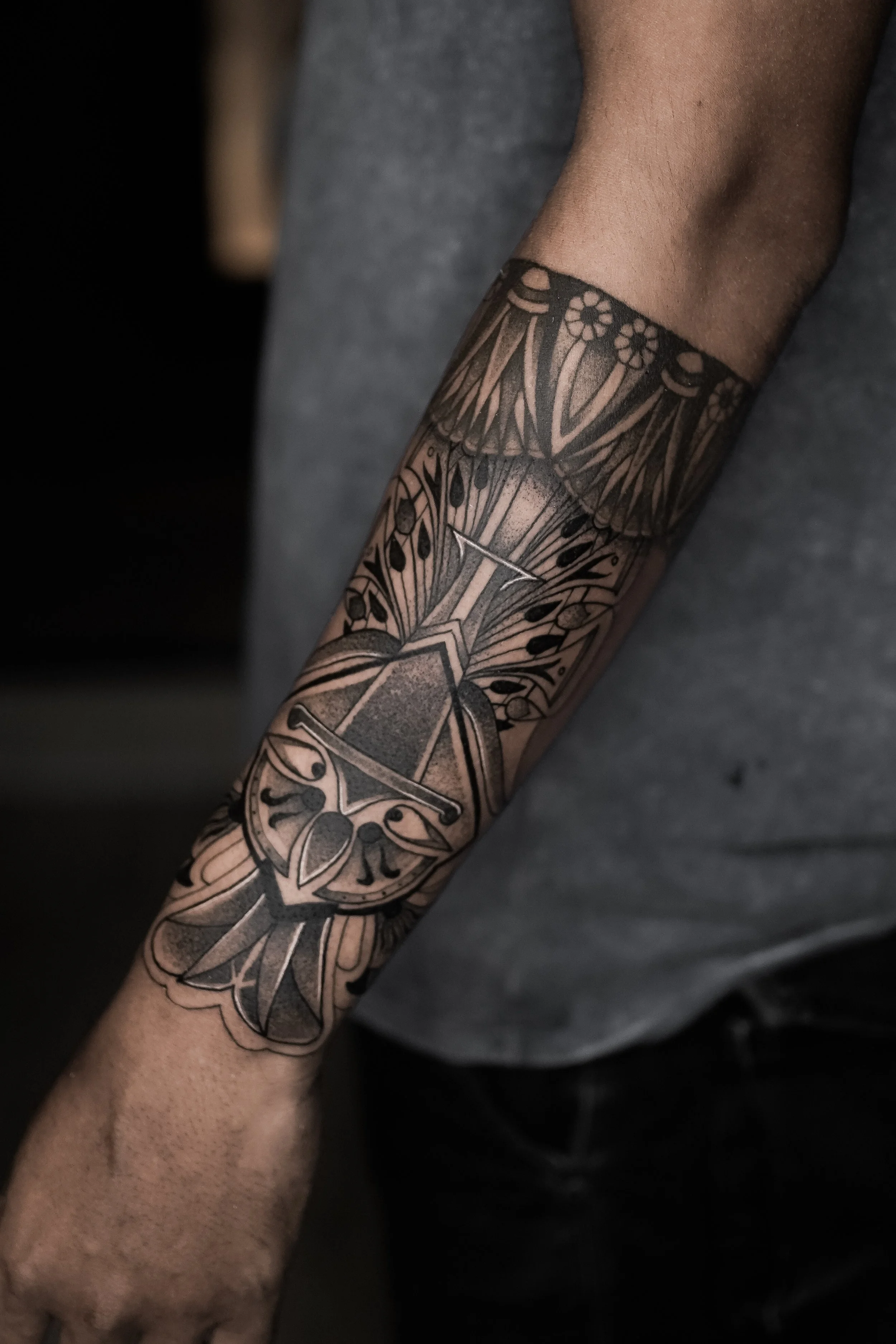 Close-up of a person's arm with a black and gray floral and geometric tattoo extending from the wrist to the elbow, wearing a gray sleeve.
