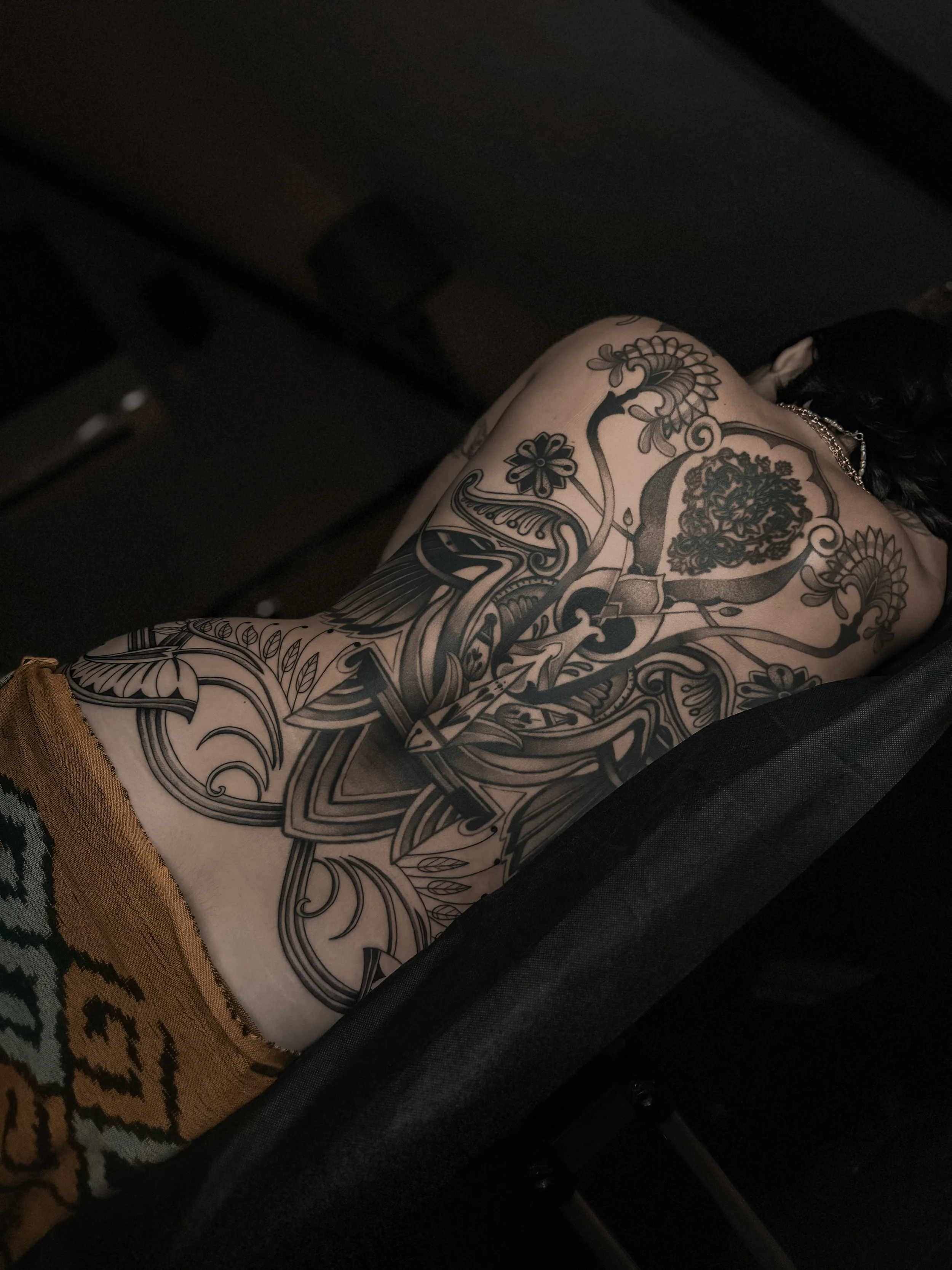 A person's arm showing intricate black and gray tattoos, including floral patterns, a snake, and decorative elements, resting on a dark surface.