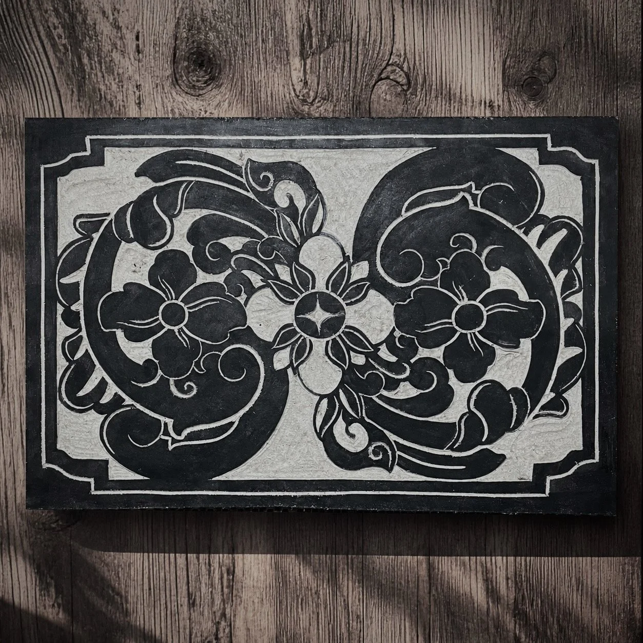 Decorative black and white floral design on a rectangular surface, possibly a metal or wood panel, with intricate swirling patterns and floral motifs surrounded by a border.