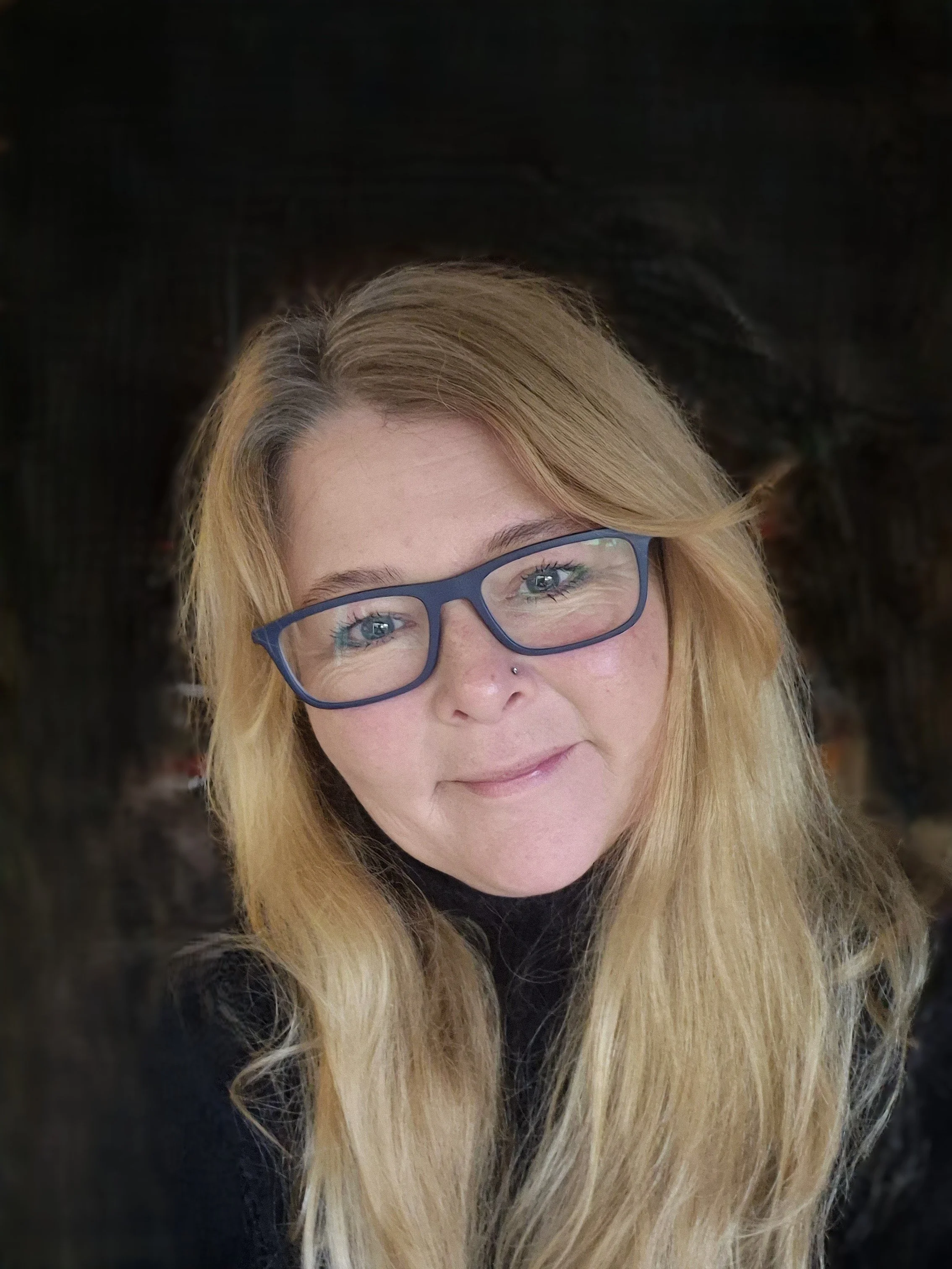 Close-up of a woman with long blonde hair, wearing blue glasses, and black clothing. She is smiling slightly and looking directly at the camera.