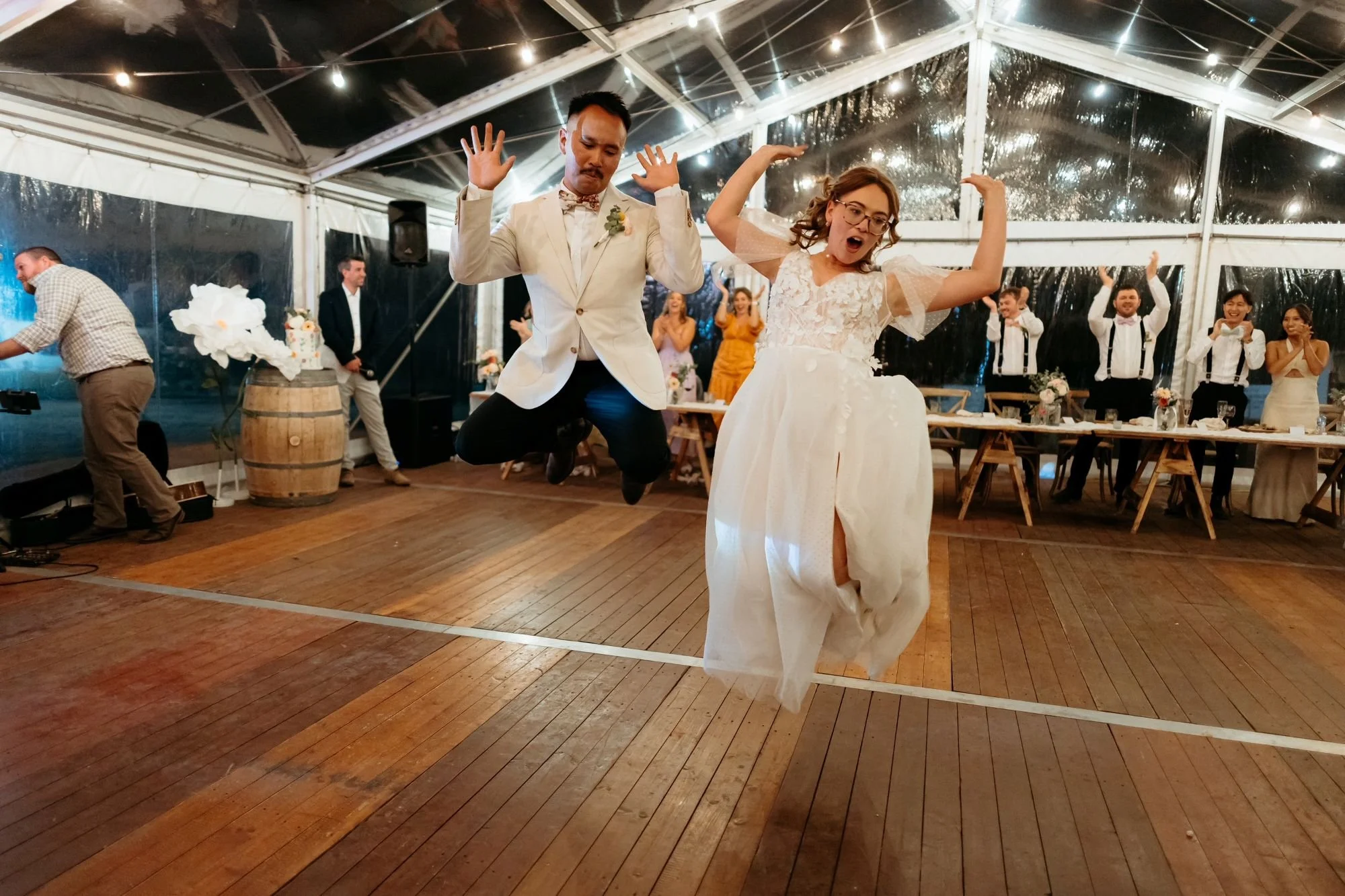 A bride and groom dancing at their wedding reception inside a tent with string lights, with guests clapping and celebrating in the background.