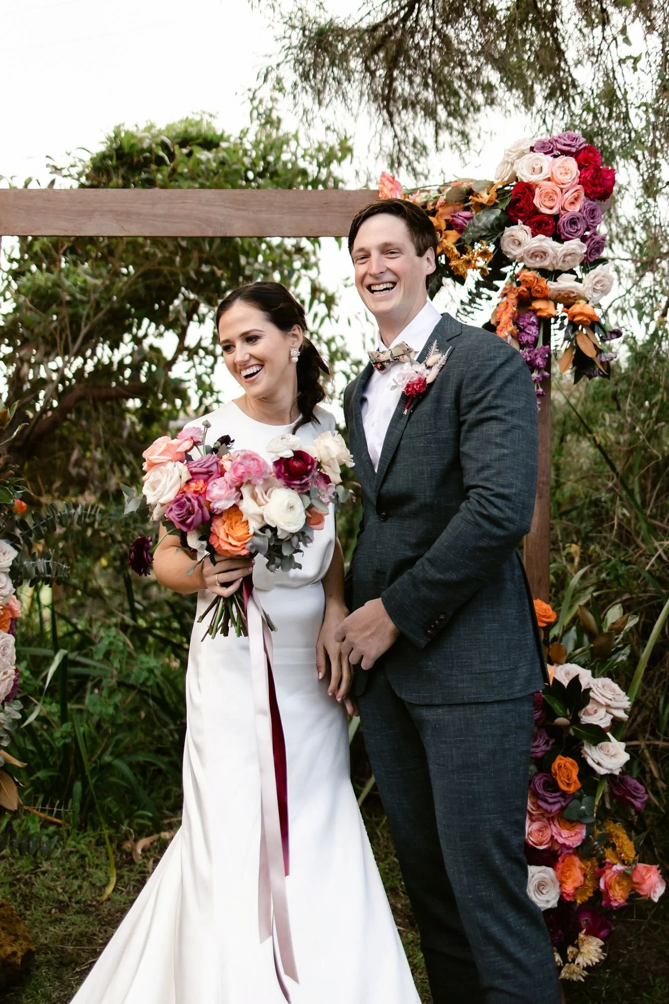 A smiling bride and groom standing outdoors in Denmark WA with a floral wedding arch behind them, holding bouquets of flowers.