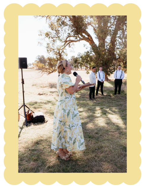 Wedding MC and celebrant Rachael Simpson in a floral dress is speaking into a microphone outdoors during a sunny day at an Albany wedding.
