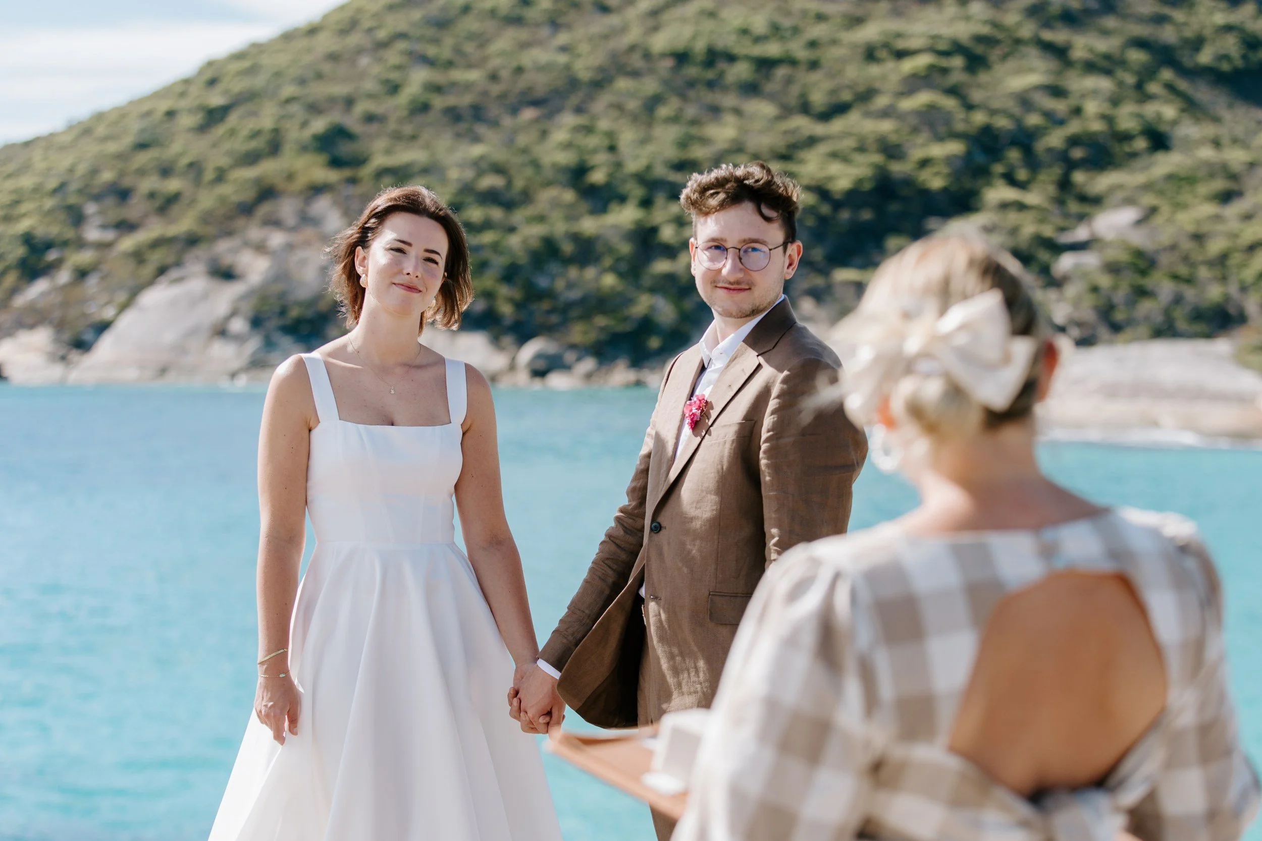 A couple dressed in wedding attire holding hands during a beach wedding ceremony in Denmark Western Australia, with a water body and green hills in the background. Rachael Simpson is the wedding officiant.