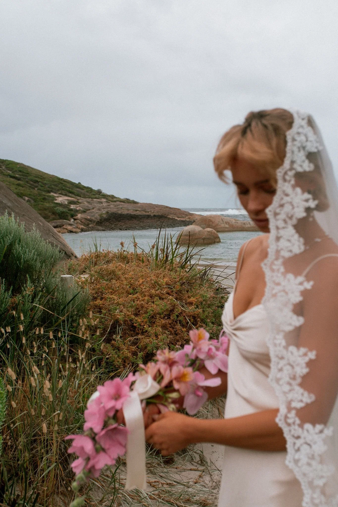 A woman in a white wedding dress with lace details, holding pink flowers, standing near a beach with rocks and greenery, overcast sky in Denmark WA.