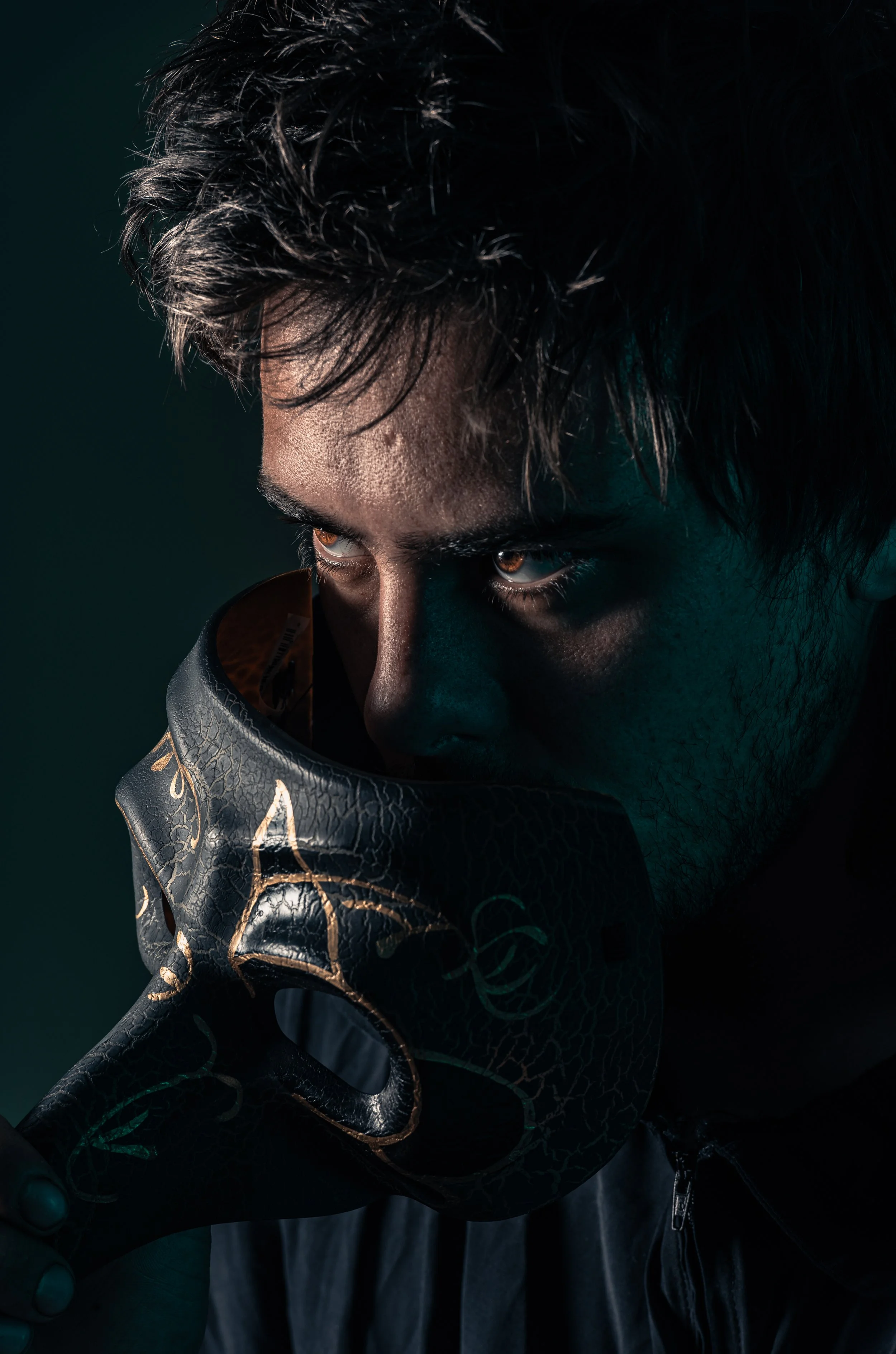 A young man with dark, textured hair and intense brown eyes, wearing a black jacket with intricate gold design, holding a black boxing glove close to his face in a dark, moody setting.