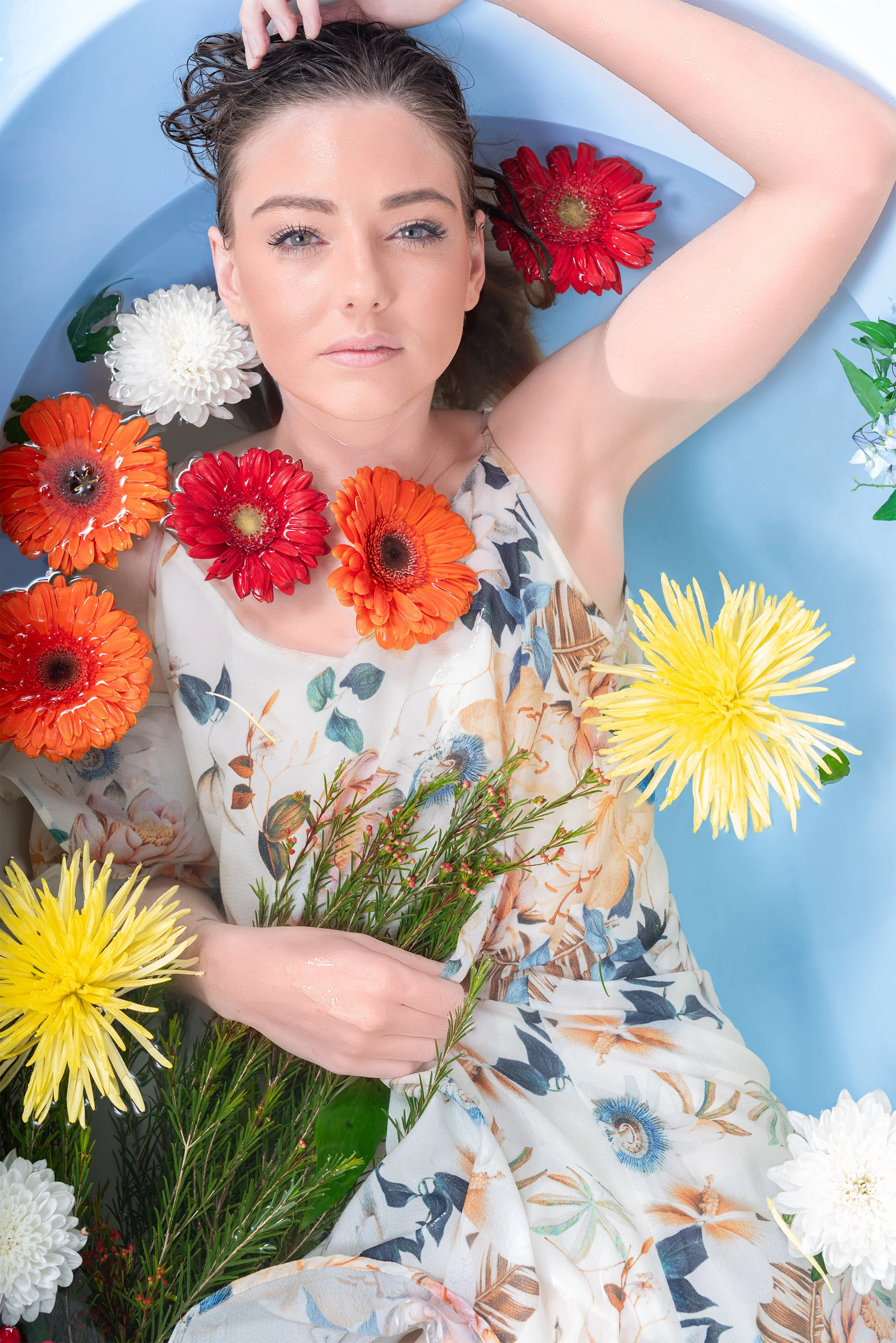 A woman with fair skin and blue eyes lying in a bathtub filled with water, surrounded by colorful flowers including red, orange, yellow, and white, wearing a floral dress and holding a bouquet of greenery.