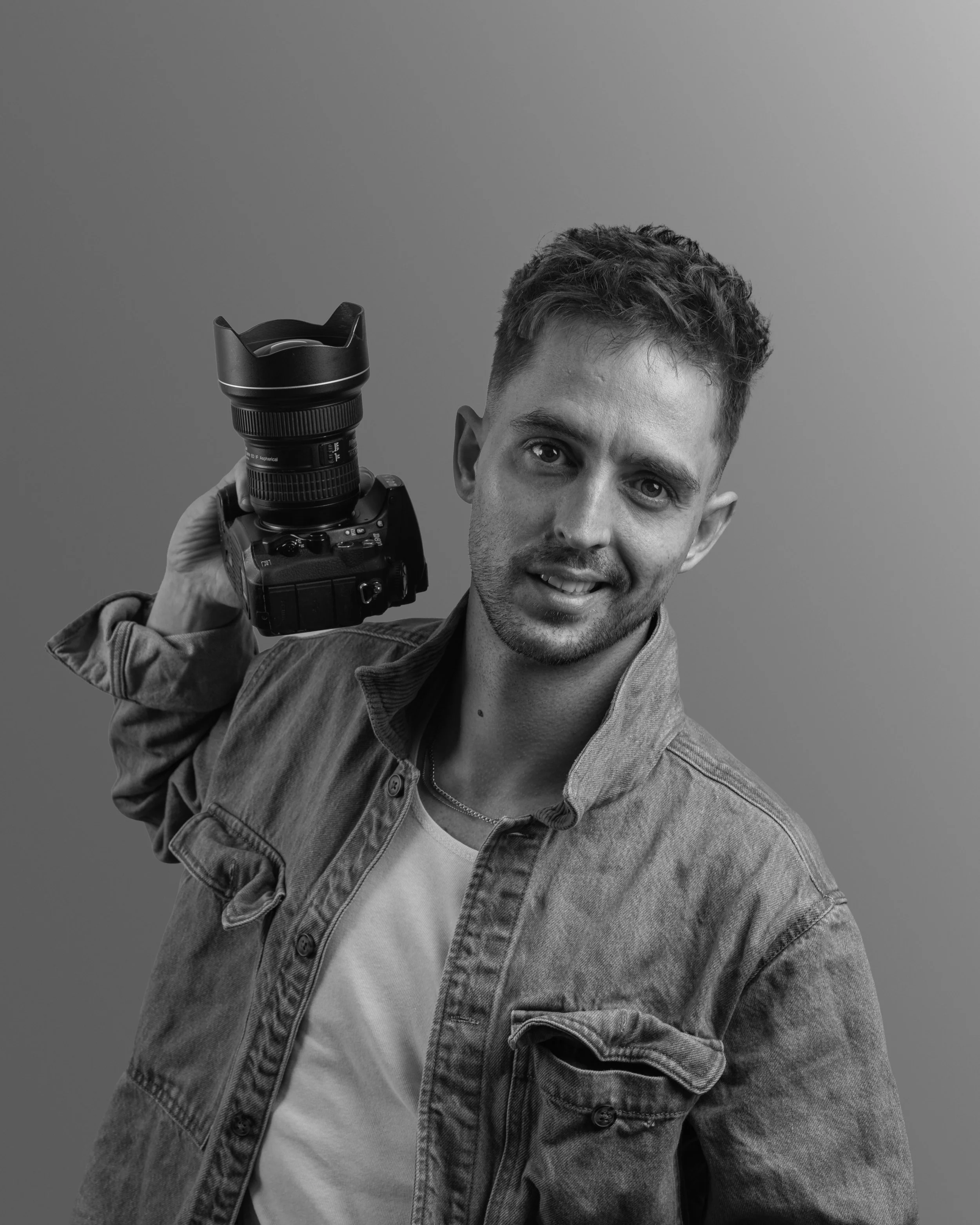A young man with short, wavy hair holding a camera with a telephoto lens. He is wearing a denim jacket over a t-shirt and is smiling at the camera against a plain background.