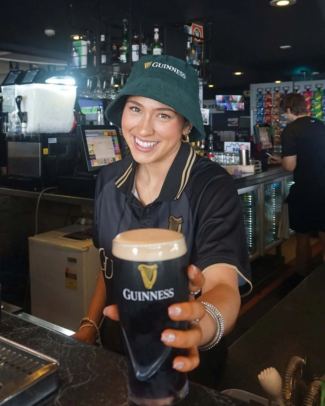 A woman smiling and holding a Guinness glass of dark beer at a bar, wearing a Guinness bucket hat and sports jersey, with bar equipment and shelves in the background.