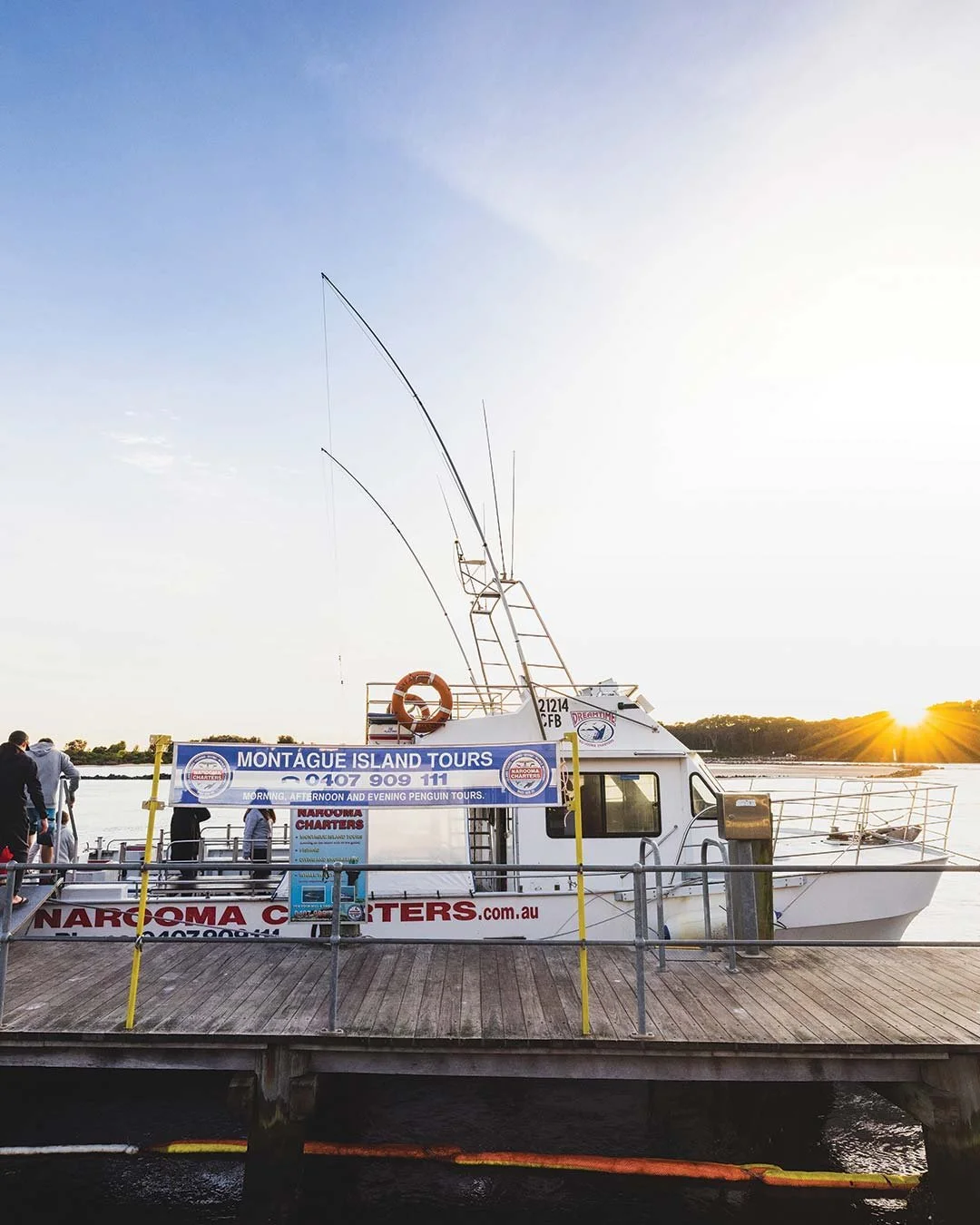 A white boat docked at a wooden pier during sunset with fishing rods on top and a sign promoting Montague Island tours.