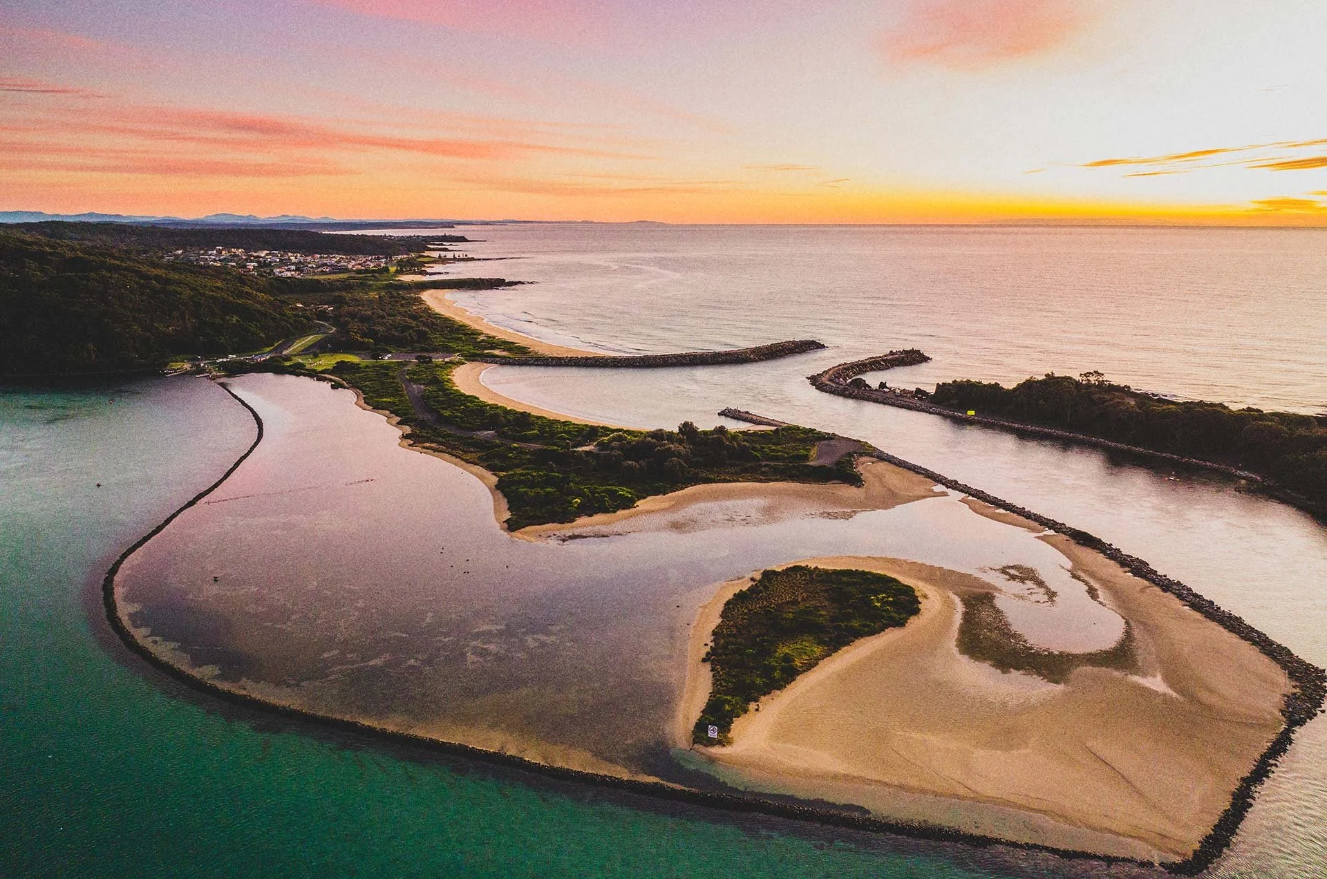 Aerial view of a coastal area at sunset, showing a river mouth with sandbars, a small beach, protective breakwaters, green vegetation, and a distant hillside.