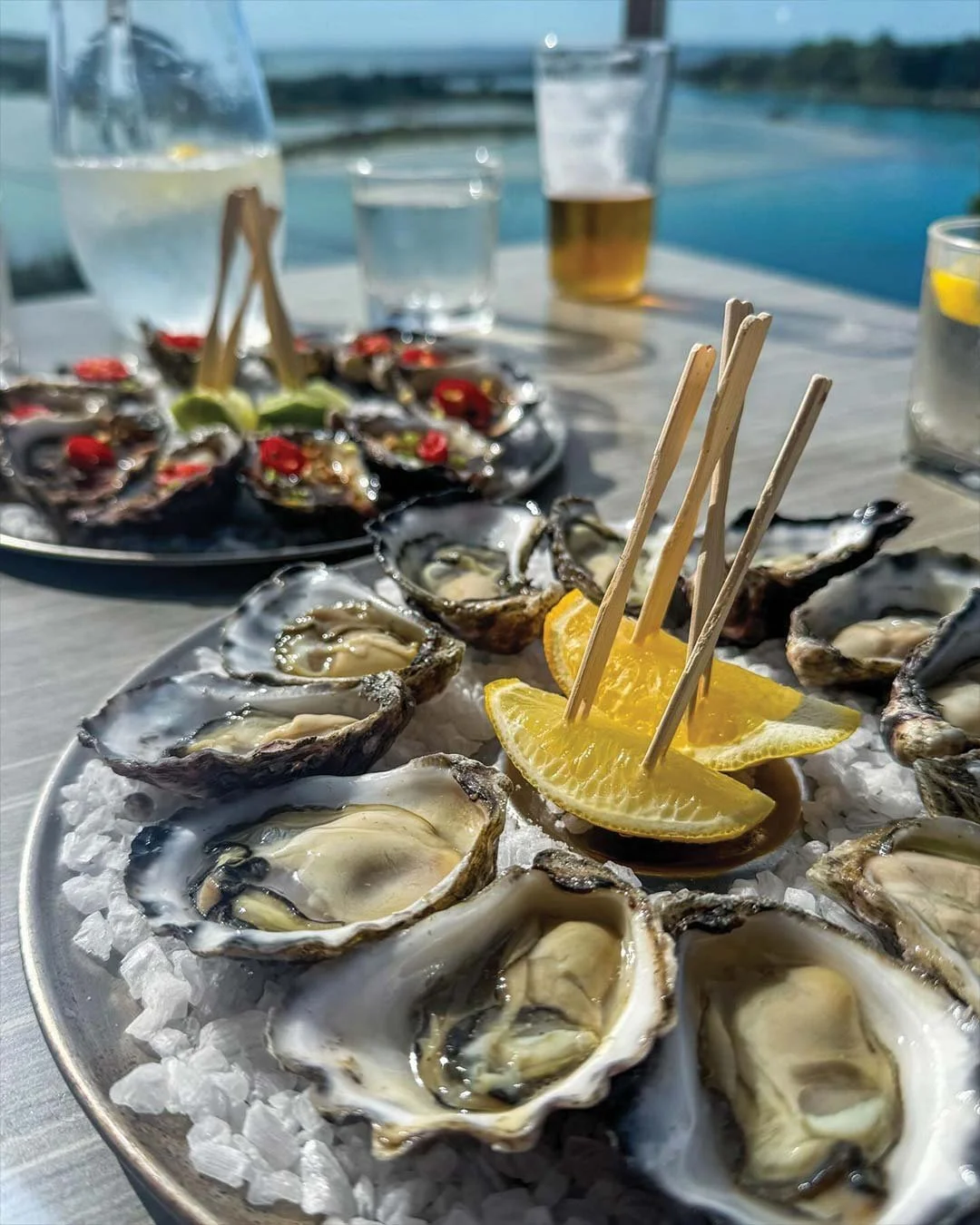A seafood platter with fresh oysters on ice garnished with lemon wedges, and another plate with raw oysters topped with red chili slices and herbs, set on an outdoor table near a body of water.