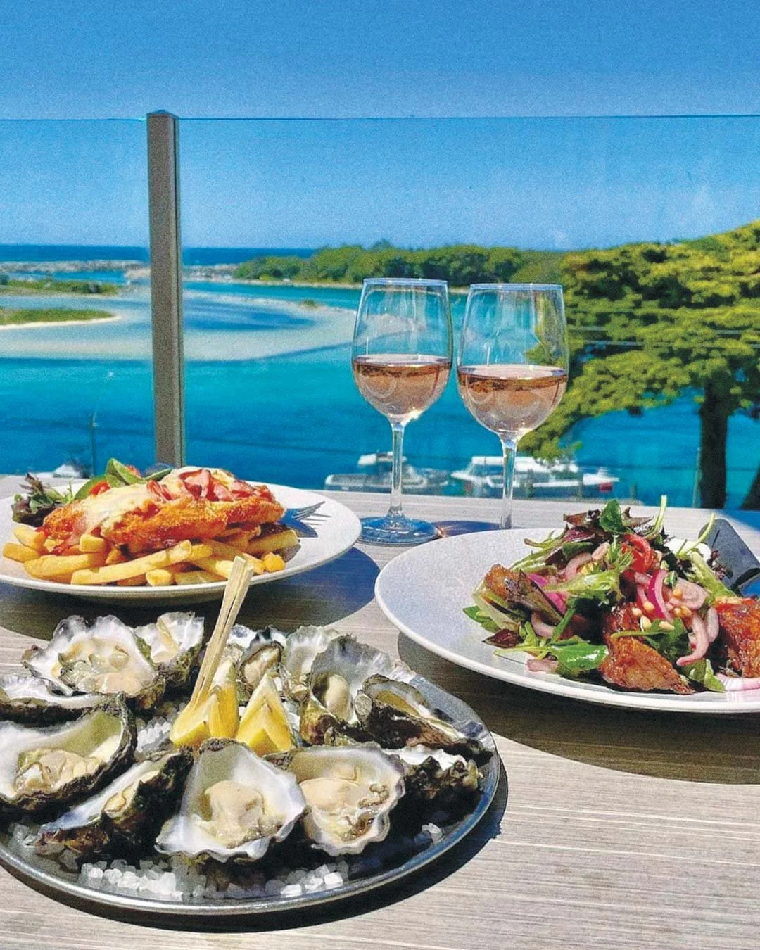 Seafood dinner with oysters, fries, fried fish with toppings, and salad on a table overlooking a waterway with boats, trees, and a blue sky.