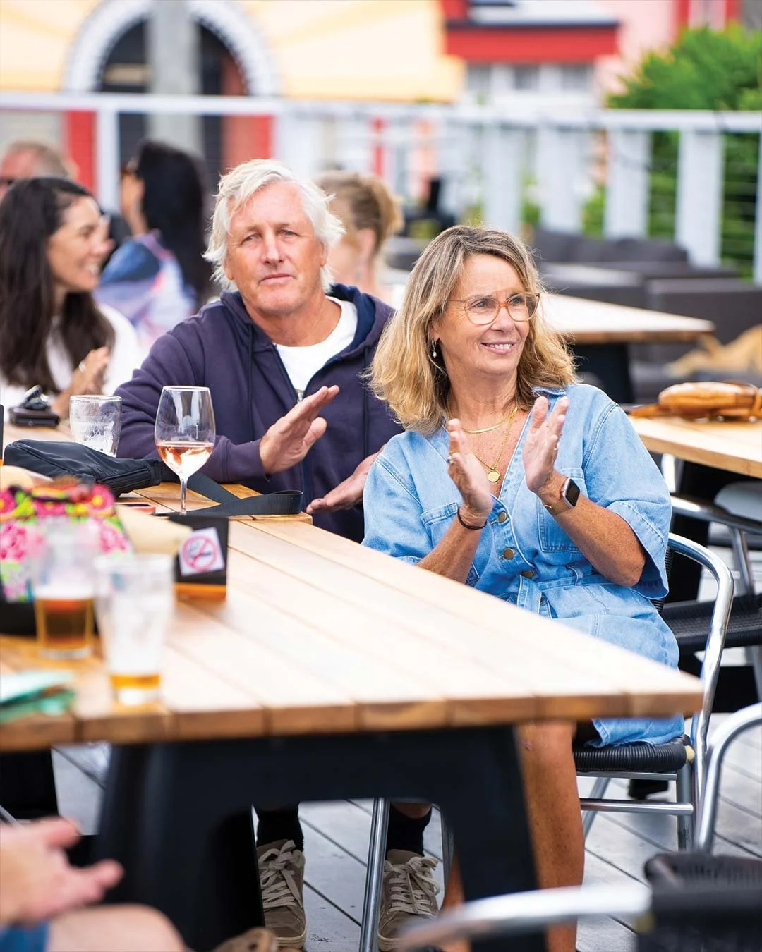 People gathered at an outdoor restaurant or gathering, some clapping and enjoying drinks, with a woman in glasses and a denim shirt smiling in the foreground.