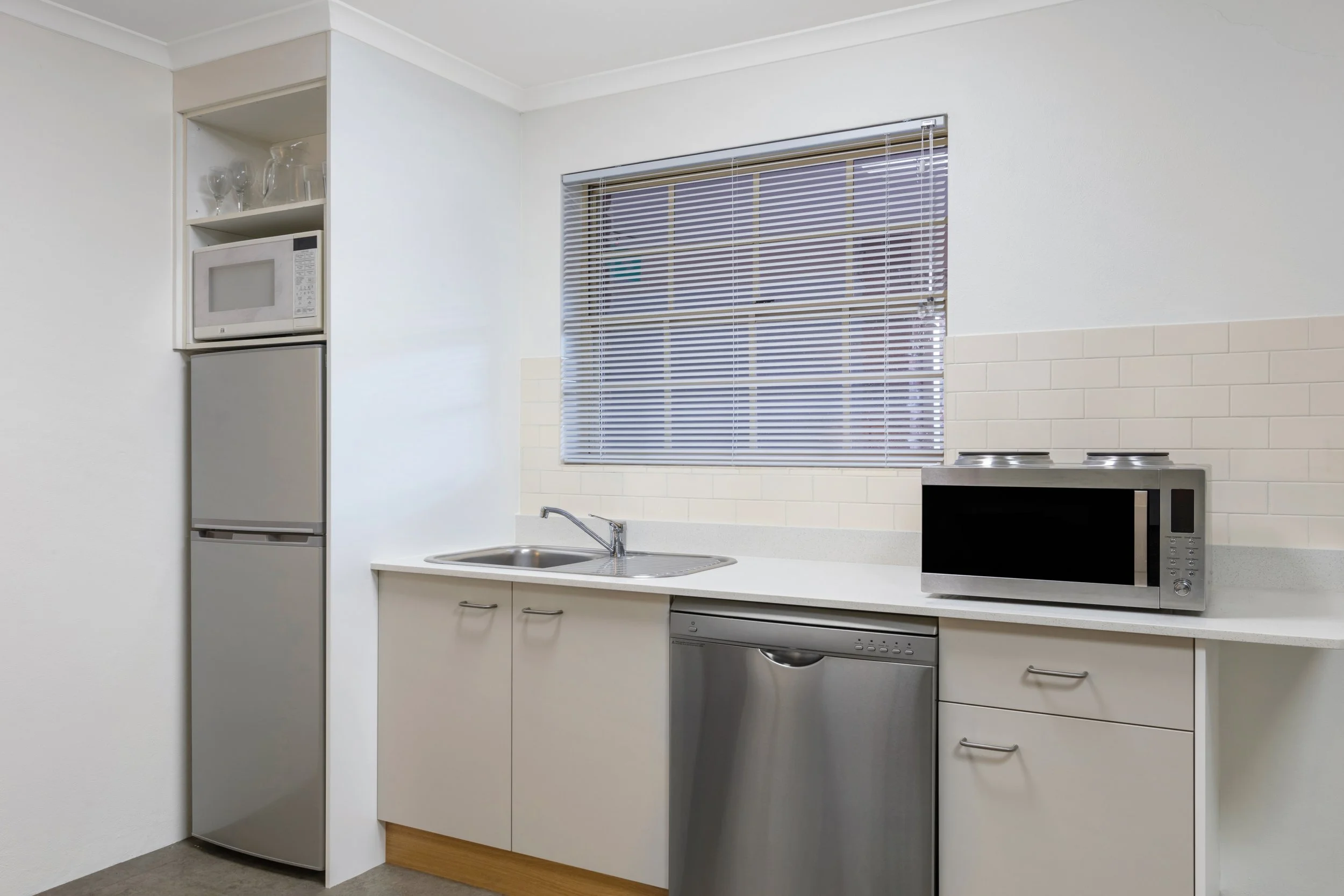 Kitchen with white walls, a window with blinds, a stainless steel sink, white cabinets, a microwave, and a stove.