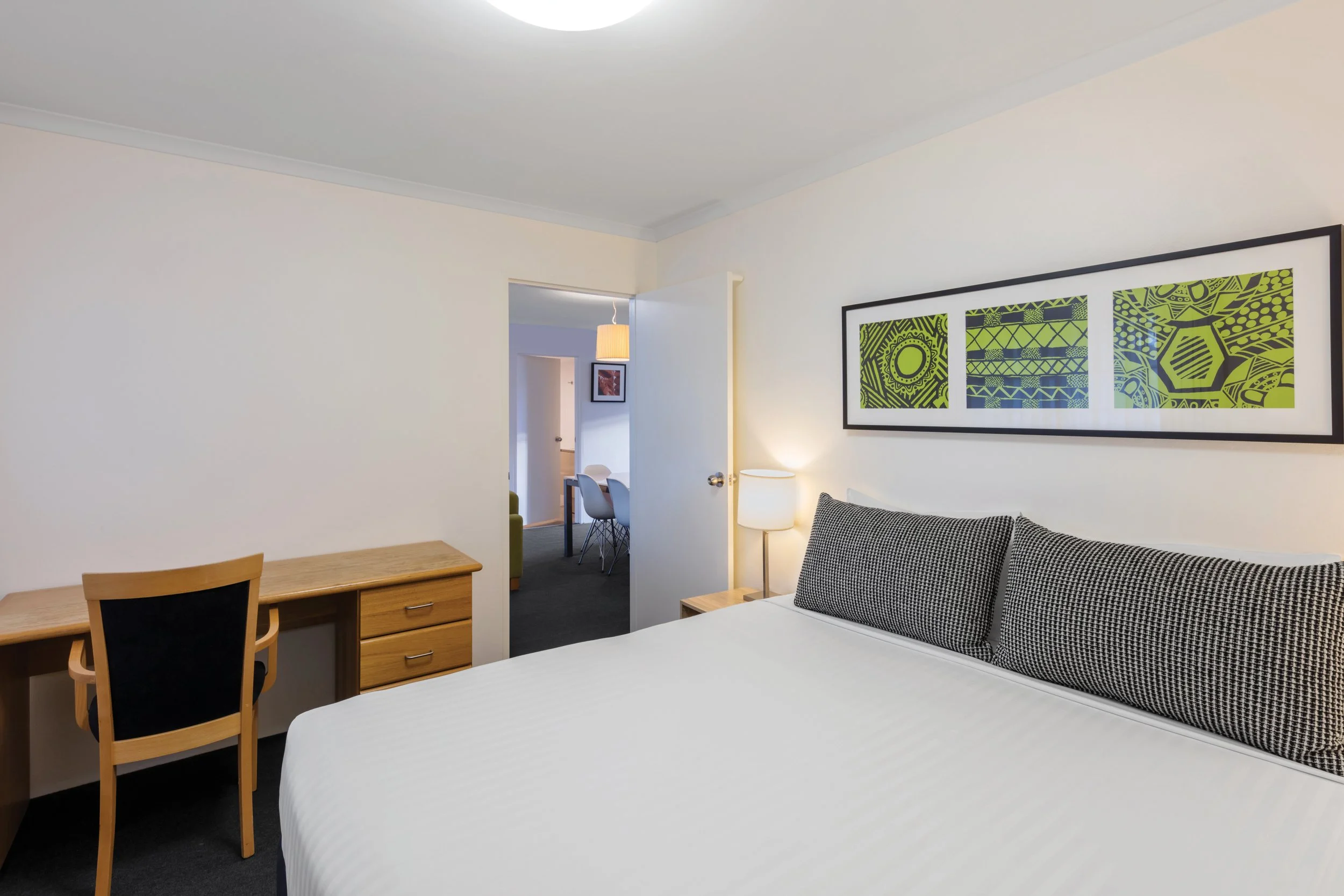 A hotel room with a bed, a wooden desk, a chair, a sofa, and wall art with green and black abstract designs.