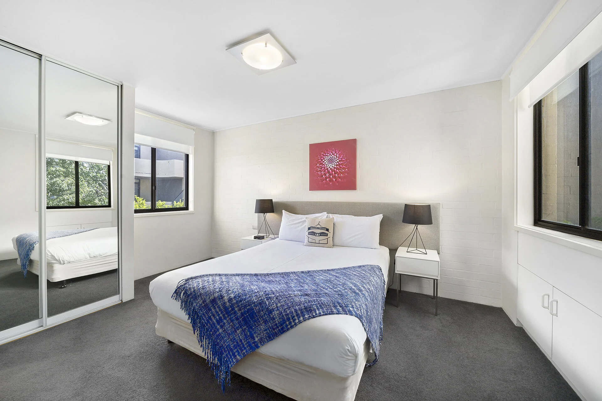 A bedroom with a bed, two nightstands with black lamps, a framed pink and white floral art piece, a white closet with mirrored sliding doors, and large windows with view of outside greenery.