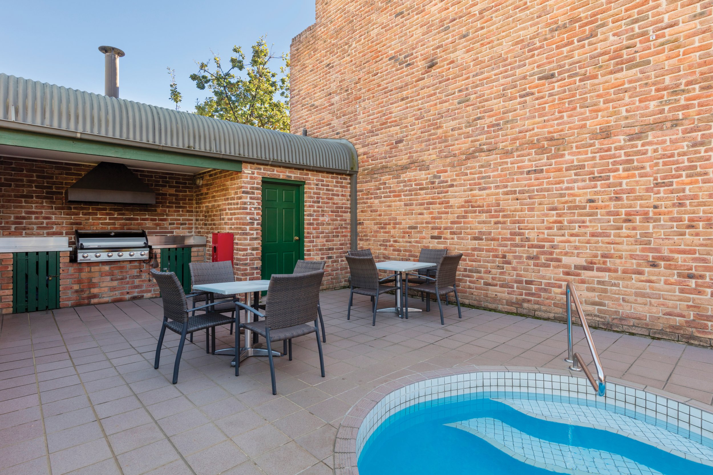 An outdoor patio area with a small swimming pool, several tables with chairs, a brick wall, a green door, a barbecue grill, and a corrugated metal roof section.