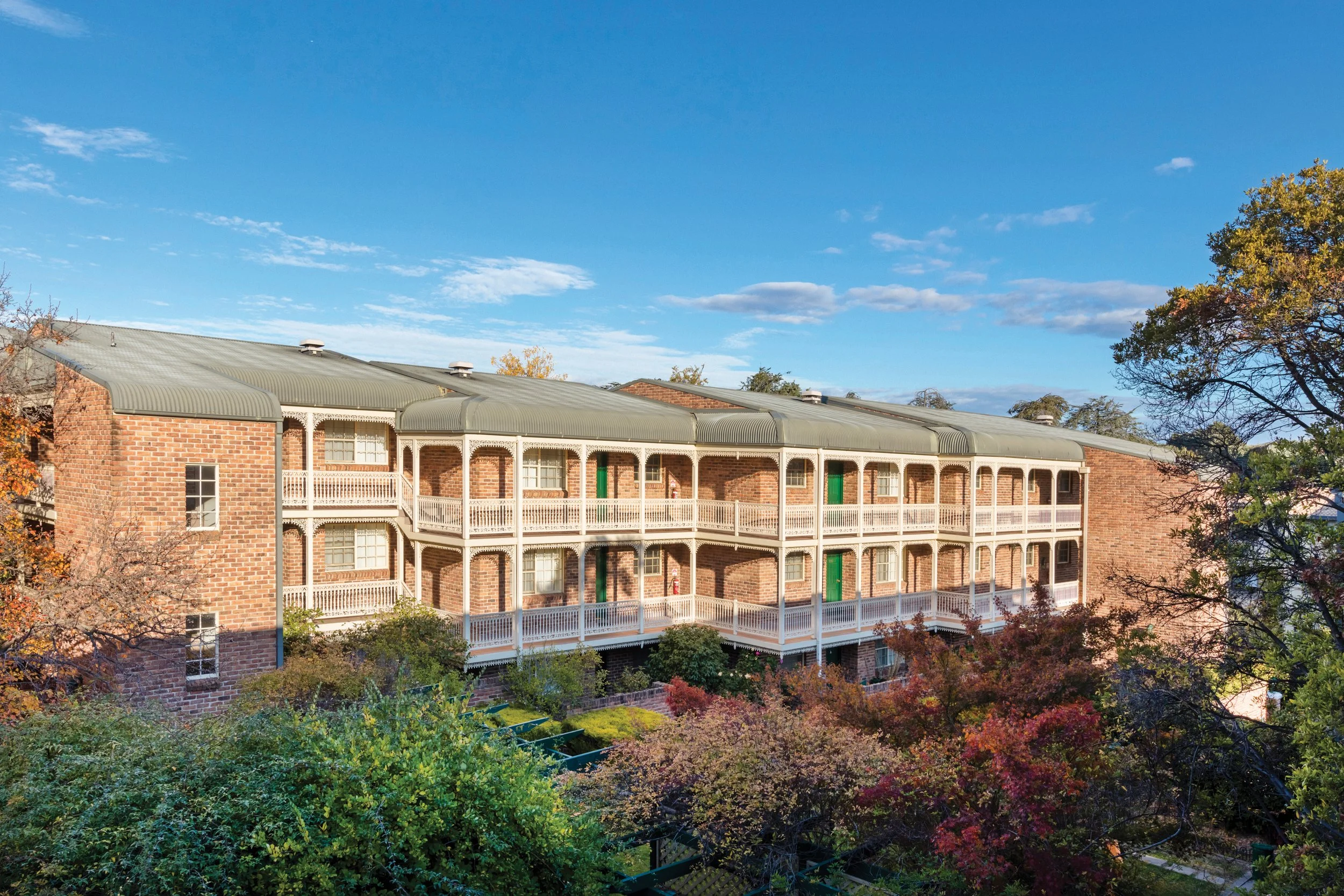 A three-story brick apartment building with white balconies under a blue sky with scattered clouds and trees with autumn foliage in the foreground.