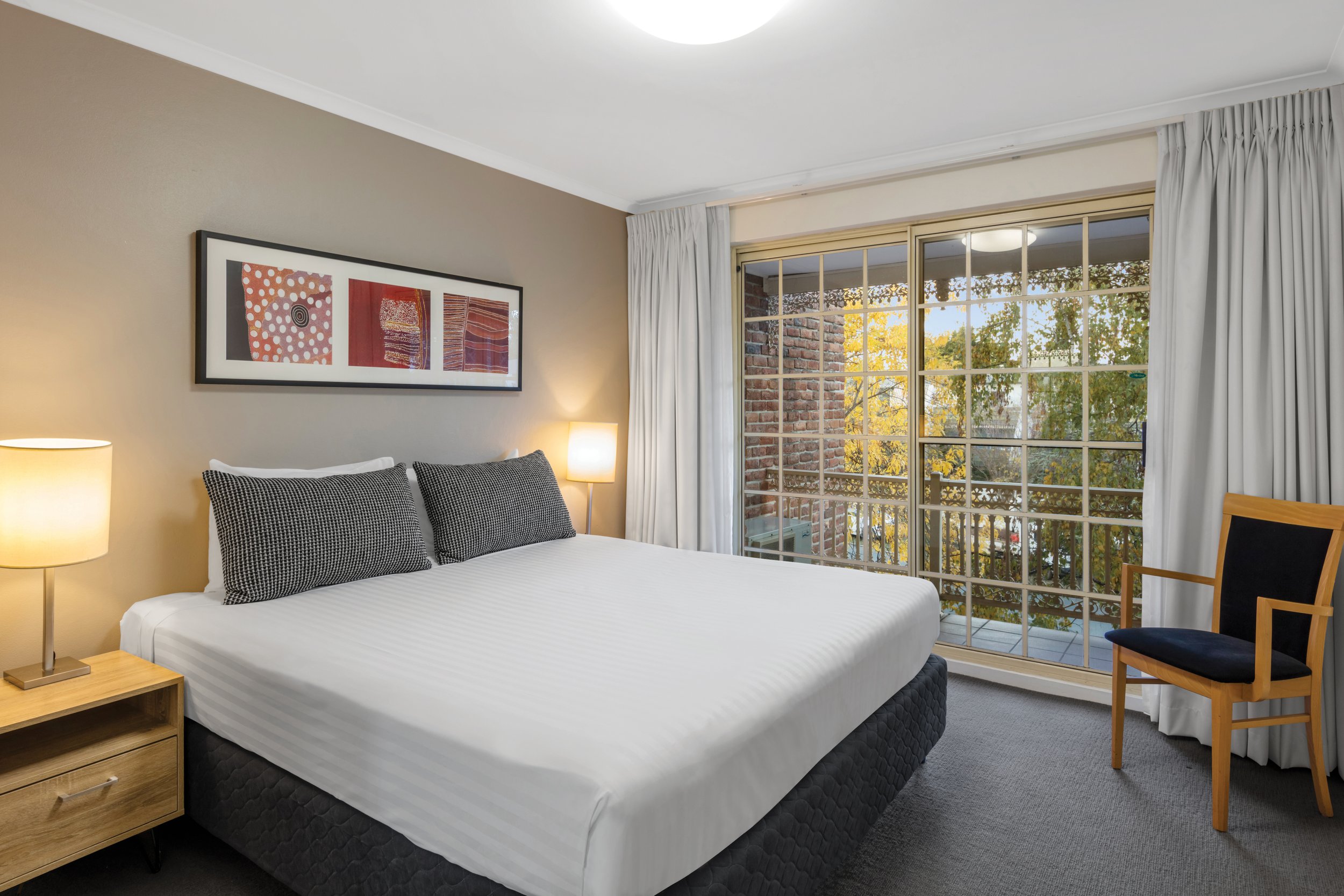 A bedroom with a white bed, two black and white checkered pillows, a wooden nightstand with a lamp, a framed abstract artwork on the wall, a large window with white curtains, a brick building outside, and part of a wooden chair.