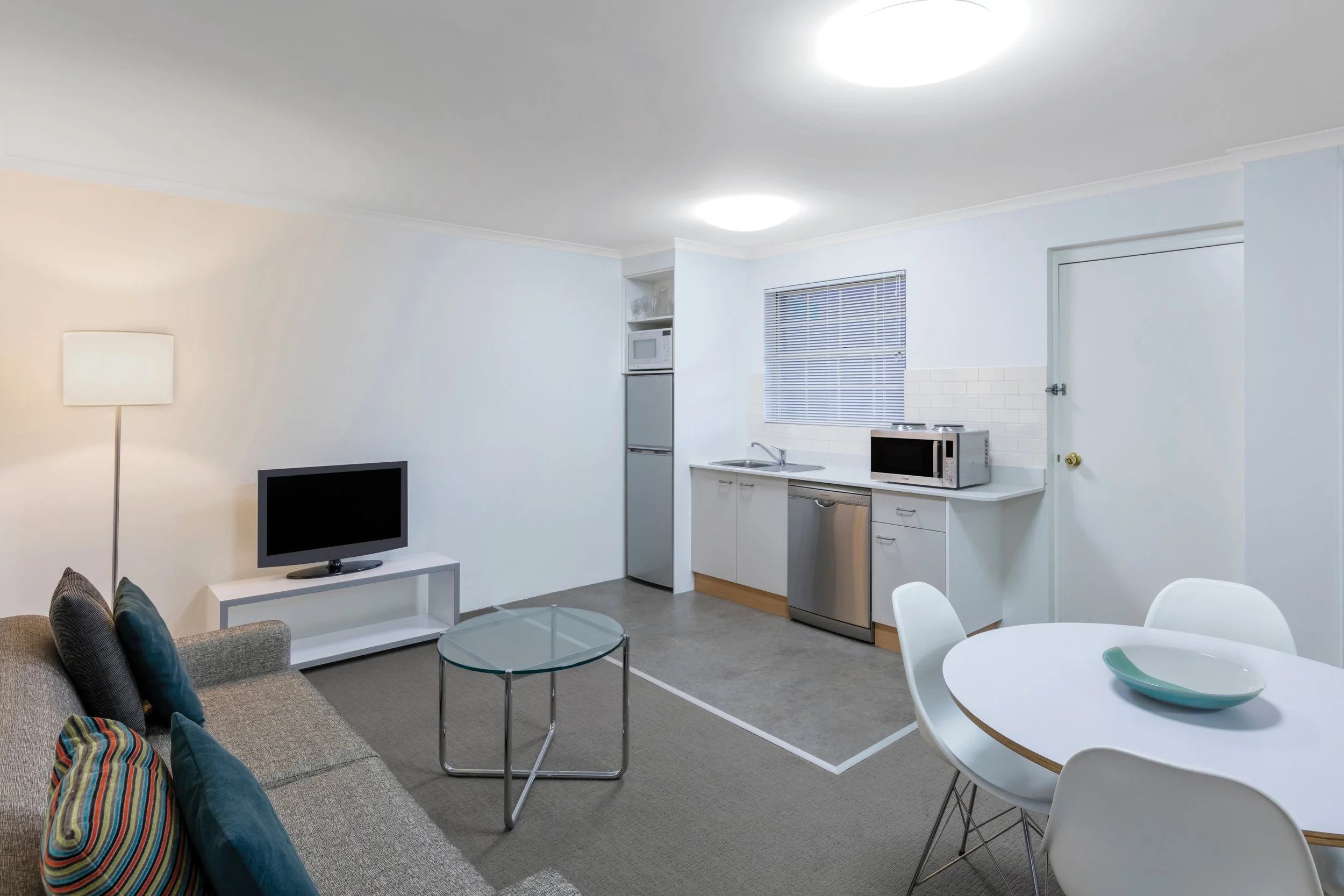 Interior view of a small apartment with a living room and kitchen area. The living room has a gray sofa with colorful pillows, a white side table, a small glass coffee table, and a floor lamp. The kitchen area includes a mini fridge, microwave, sink,