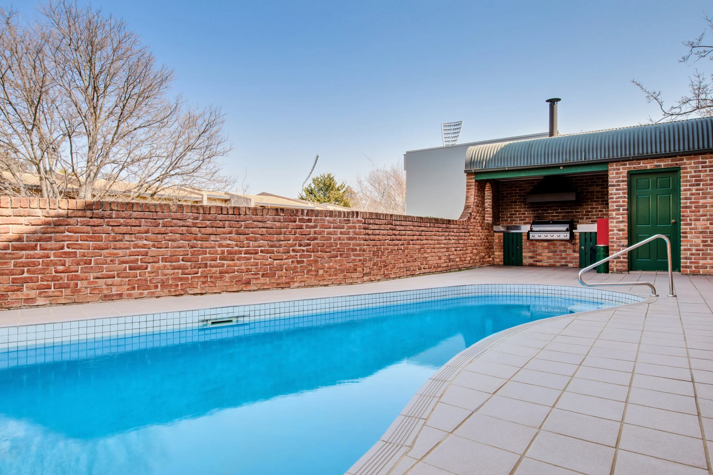 A backyard swimming pool with blue water, surrounded by beige tiles, next to a red brick wall and a small outdoor kitchen area with a grill, green door, and metal roof. Leafless trees and a clear blue sky are in the background.