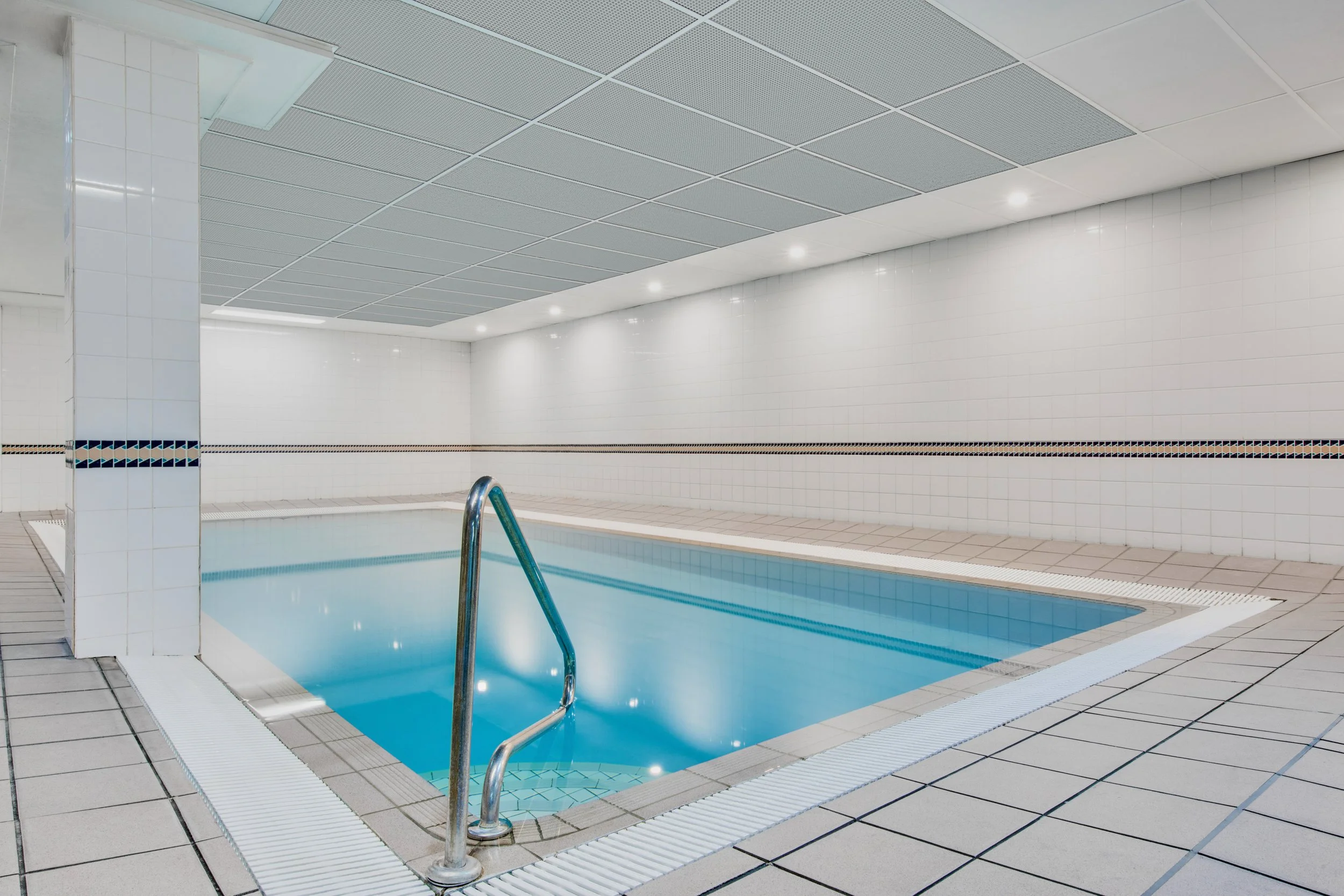 Empty indoor swimming pool with ladder, white tiled walls, and ceiling with fluorescent lights.