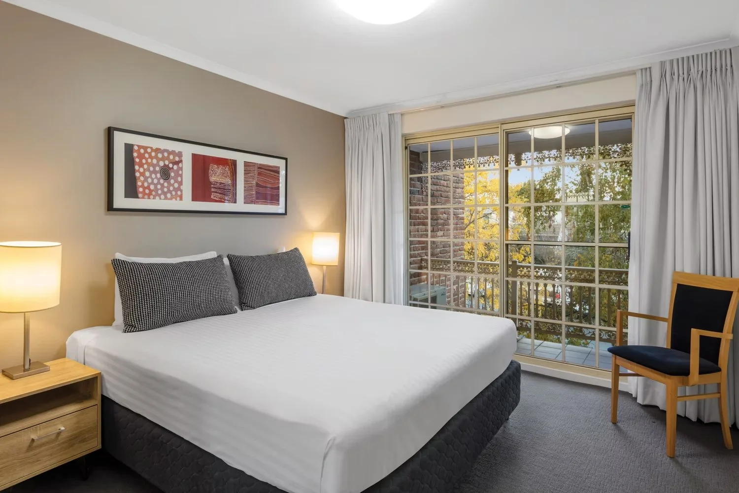 A bedroom with a large bed, two pillows, nightstands with lamps, abstract wall art, and a large window with white curtains showing trees outside.