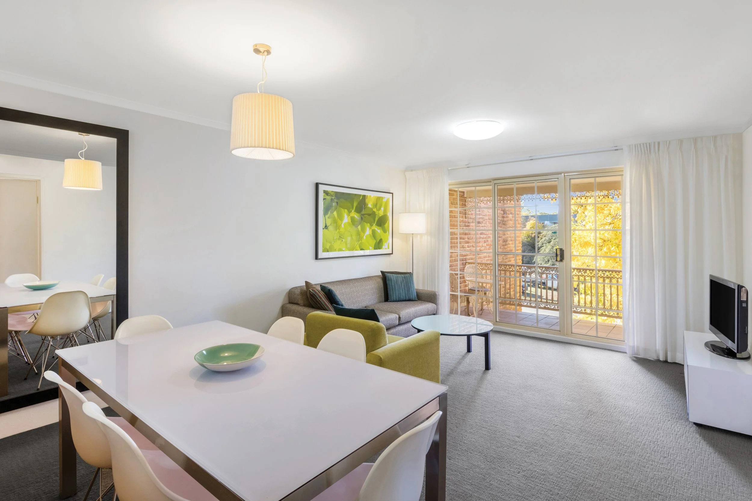 Bright living room and dining area with large window showing autumn trees, modern white dining table with chairs, beige sofa with pillows, artwork on wall, ceiling lights, and a balcony visible outside.