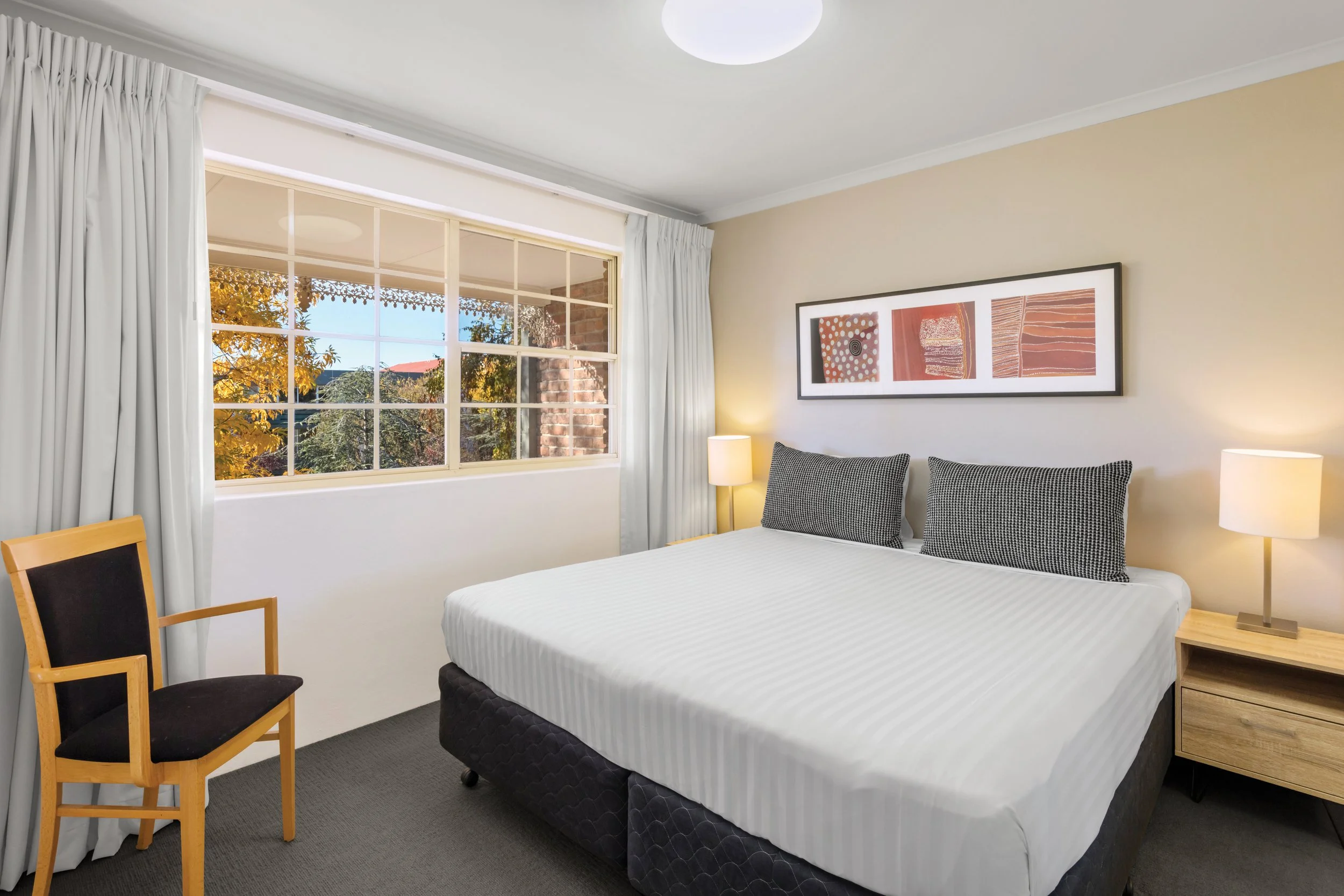 A bedroom with a window showing trees outside with orange and green leaves, a bed with a white and black patterned headboard and white bedding, framed artwork on the wall, a nightstand with a lamp, and two chairs, one wooden with a black cushioned se