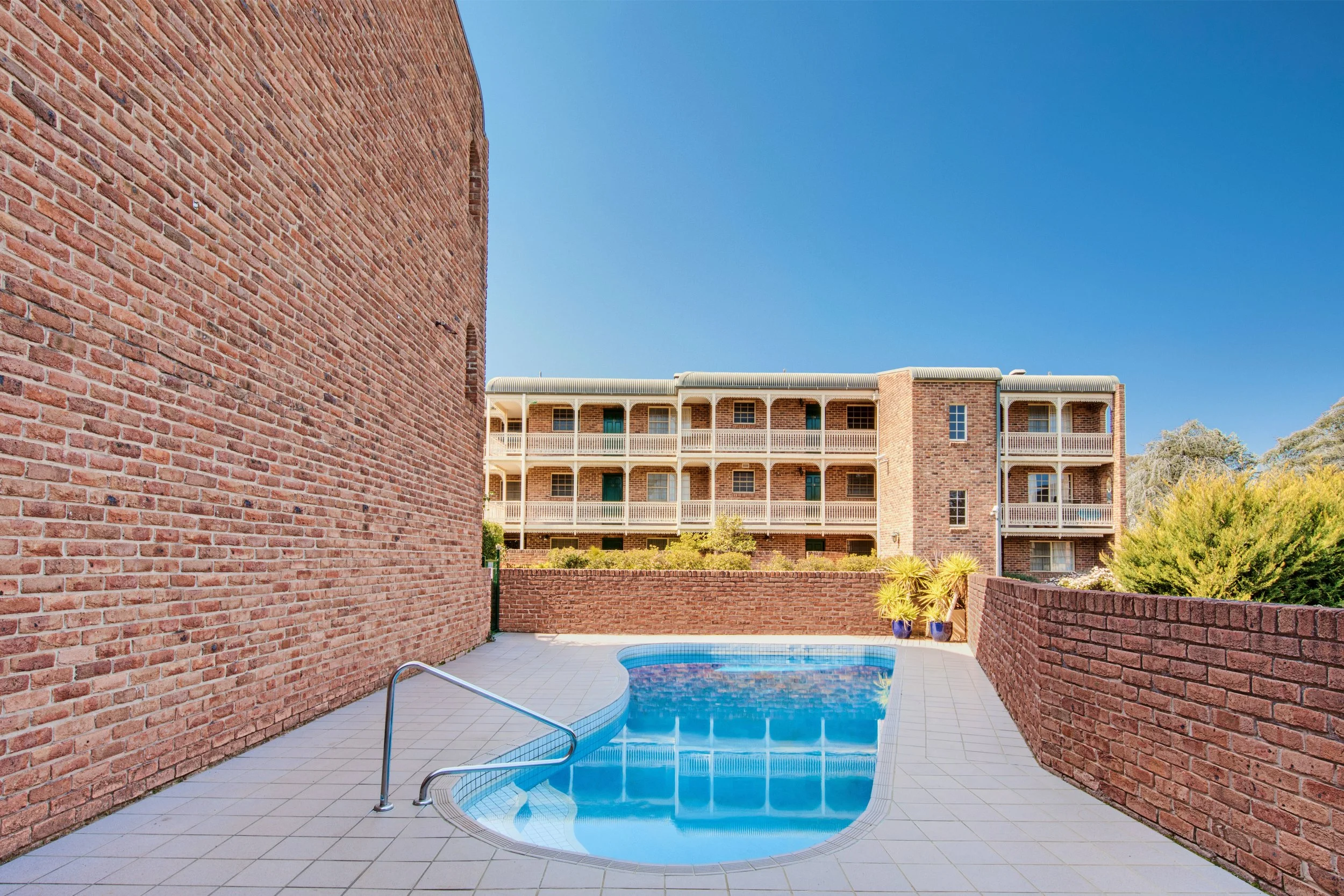 A small round swimming pool with a metal handrail located in a concrete patio area, enclosed by red brick walls and apartment buildings in the background.
