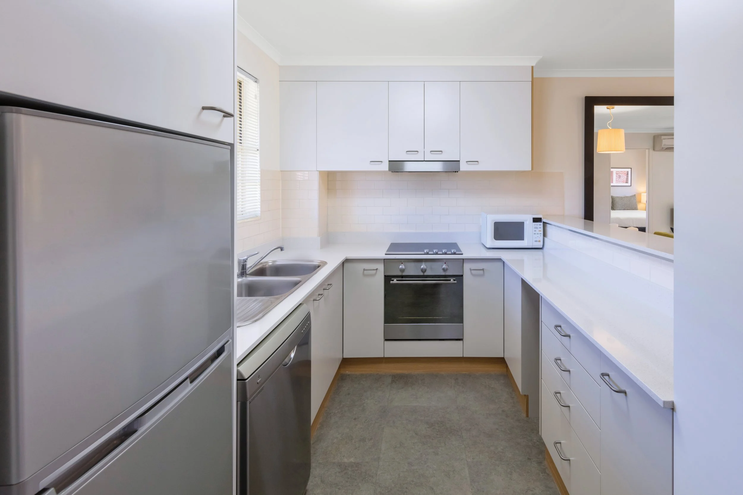 A modern, minimalist kitchen with white cabinets, a stainless steel oven, microwave, refrigerator, and a double sink. There is a window with blinds, and an opening to a living room visible with a hanging light fixture and picture frame.