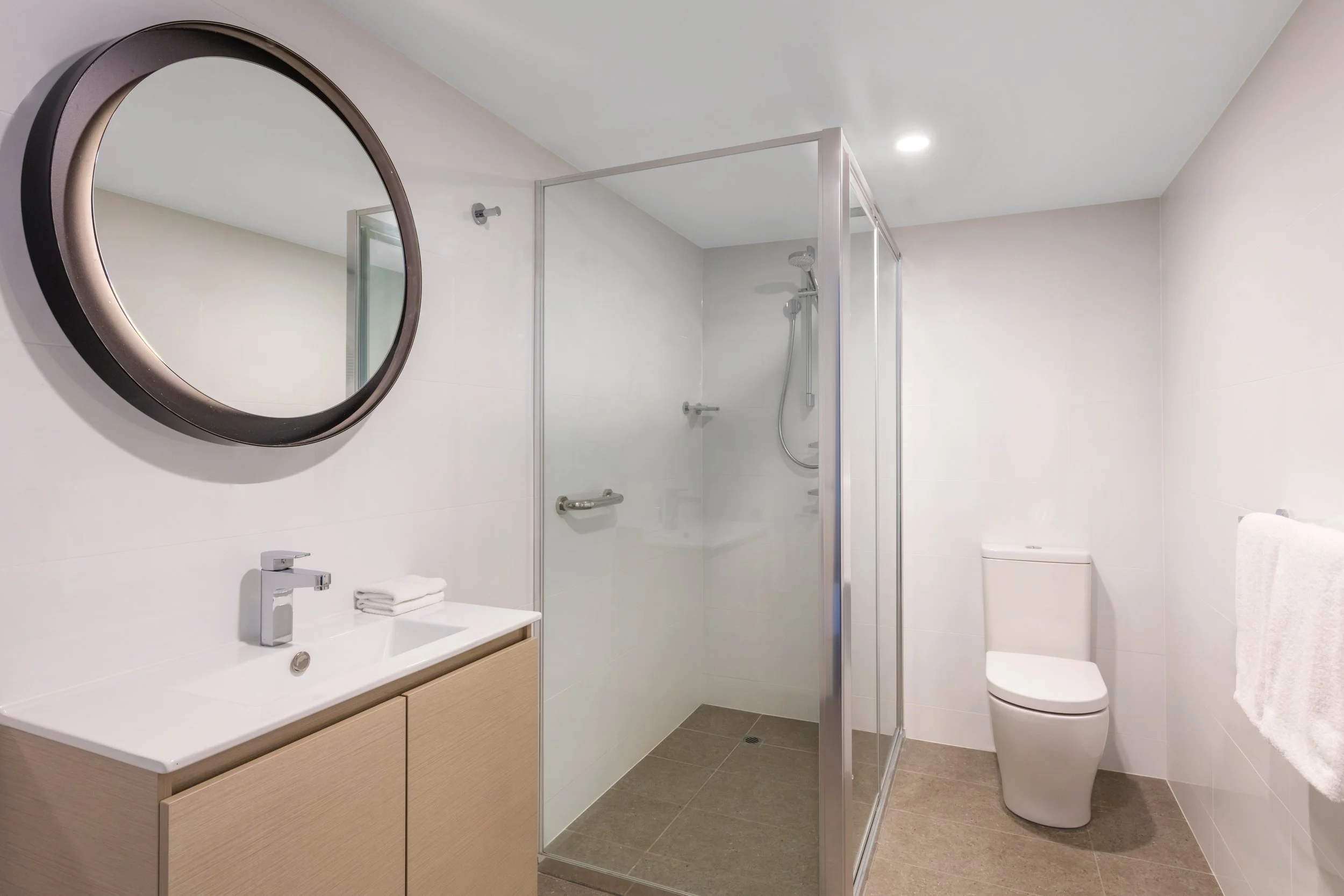 A modern bathroom featuring a round mirror above a white sink with a wooden cabinet. To the right, there is a toilet and a walk-in shower enclosed with a glass door. The bathroom has white walls, a beige tile floor, and a white ceiling with recessed 