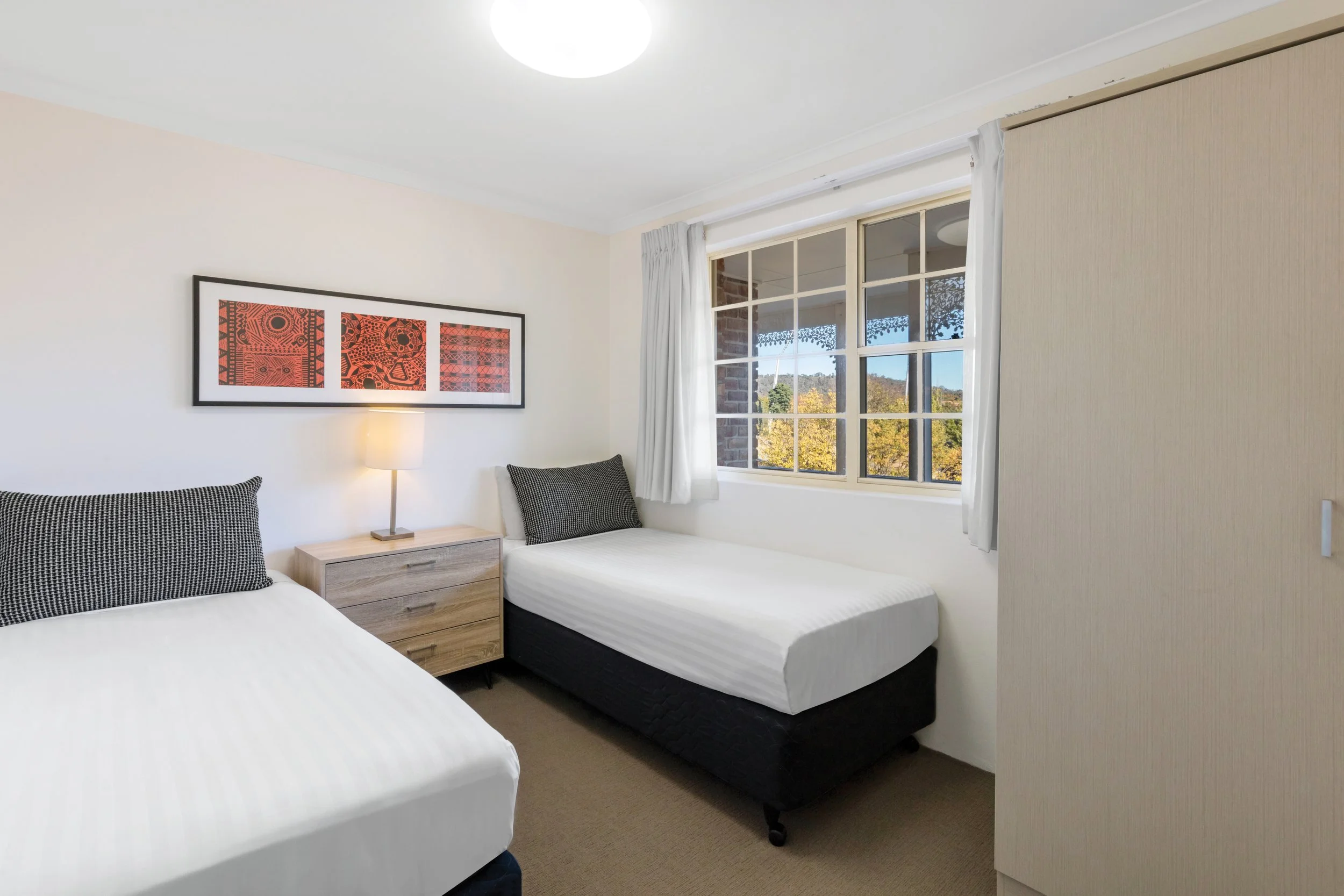 Interior view of a bedroom with two twin beds, a nightstand, a lamp, a framed artwork with tribal patterns, a window with white curtains, and a view of trees and sky outside.
