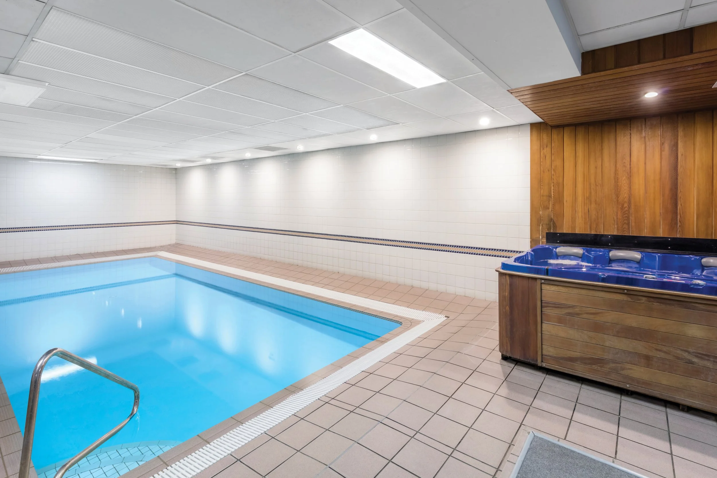 Indoor swimming pool with blue water, surrounded by beige and white tiled flooring and white tiled walls, with a wooden hot tub in the background.
