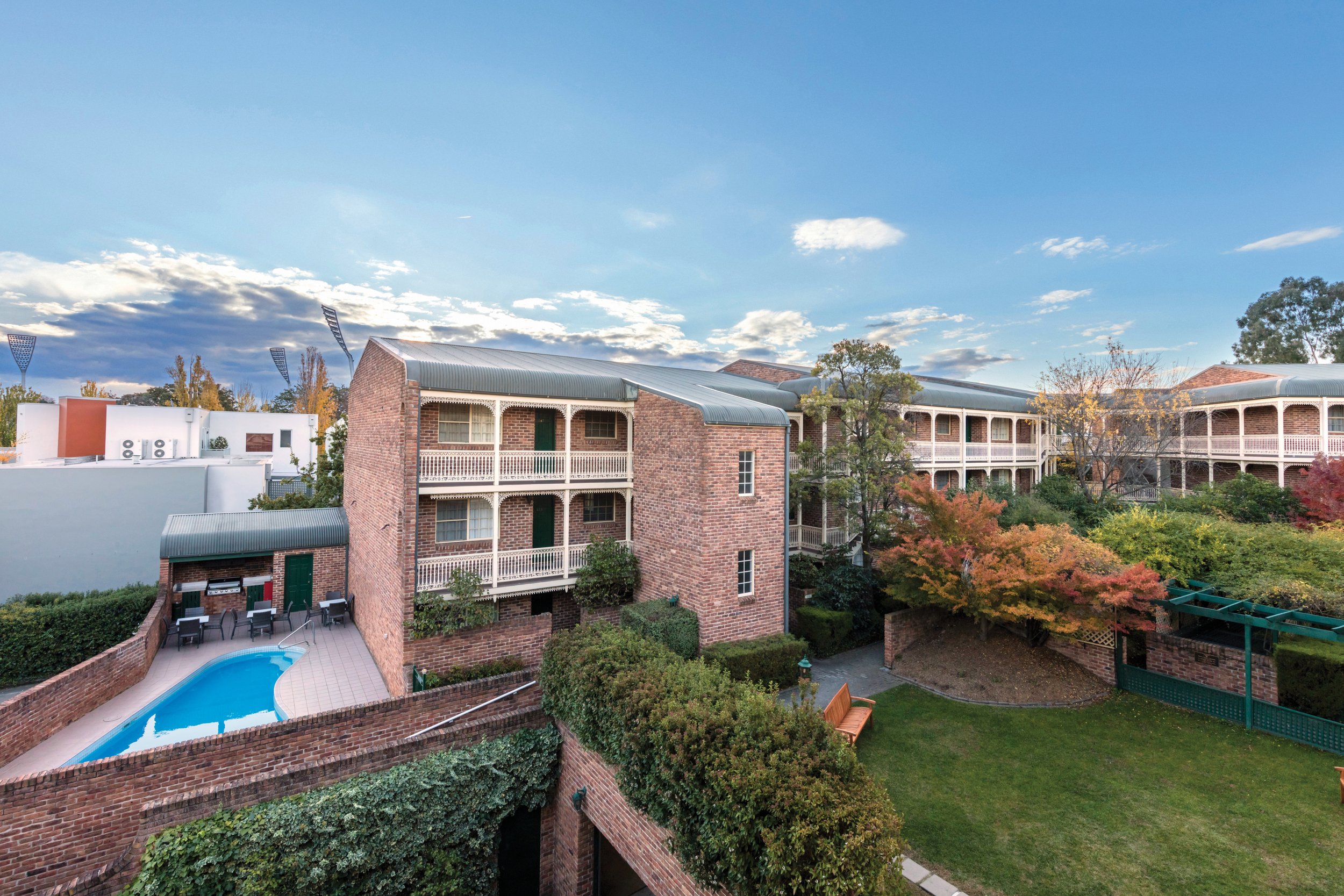 Multi-story brick residential buildings with white balconies, a swimming pool, outdoor seating area, surrounded by trees with fall foliage, under a partly cloudy sky.