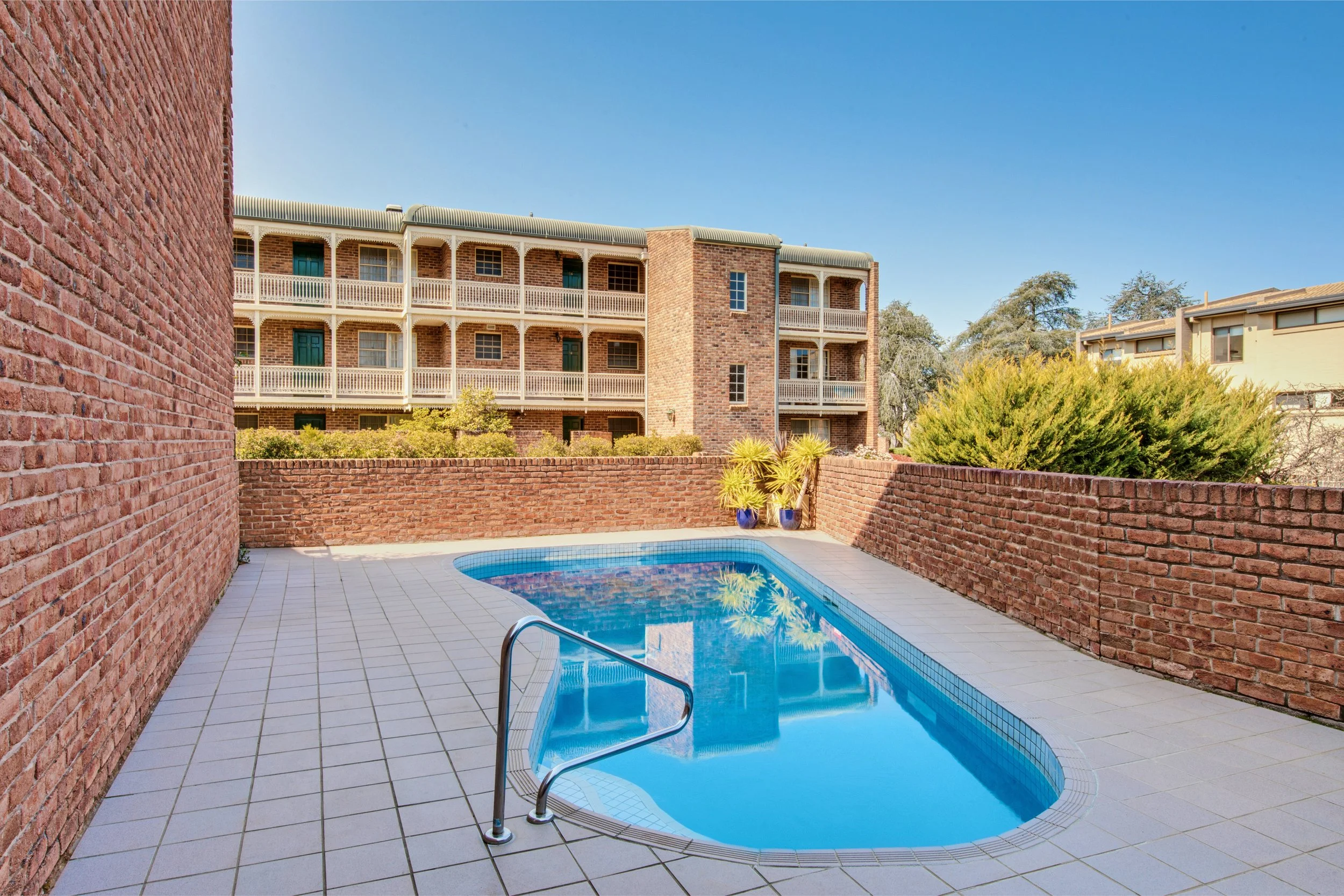A residential apartment complex with multiple story brick buildings and a backyard swimming pool area with a brick wall, potted plants, and trees under a clear blue sky.
