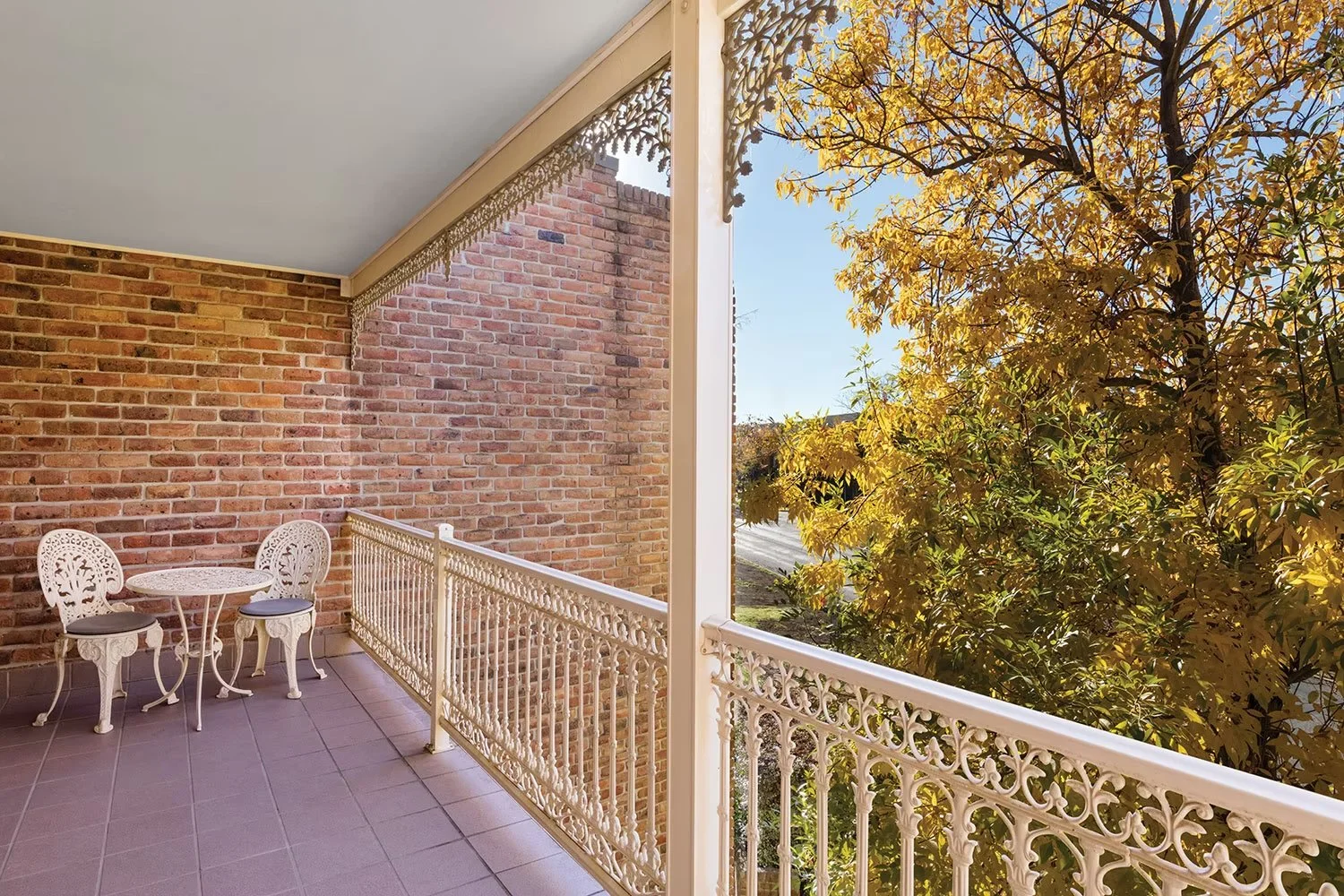 Balcony with white ornate railing, white patio furniture with two chairs and a small round table, brick wall to the left, overhanging ceiling, large tree with yellow leaves outside, clear blue sky, and a street visible in the background.
