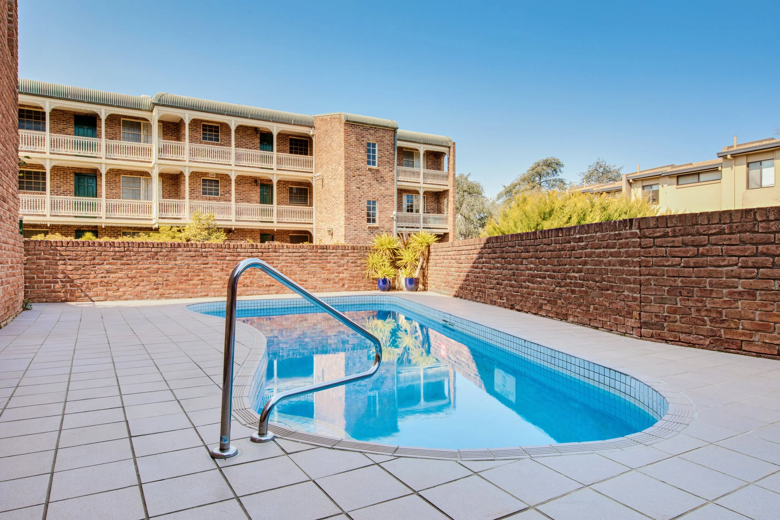 Empty swimming pool with a metal handrail, surrounded by white tiled deck and brick walls, with multi-story brick and light-colored residential buildings in the background under a clear blue sky.