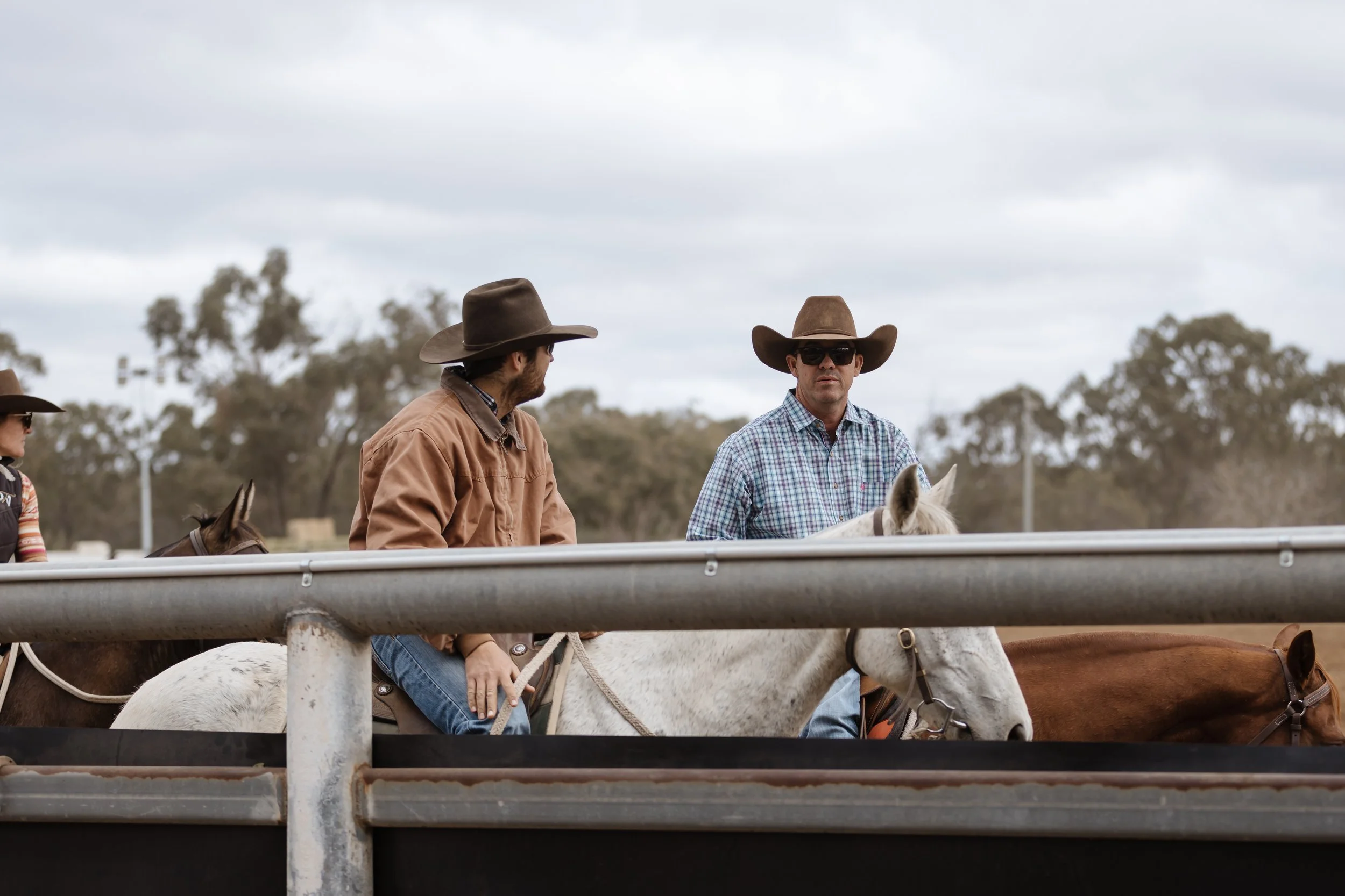 Three people on horseback wearing cowboy hats, sitting behind a metal railing outdoors on a cloudy day.