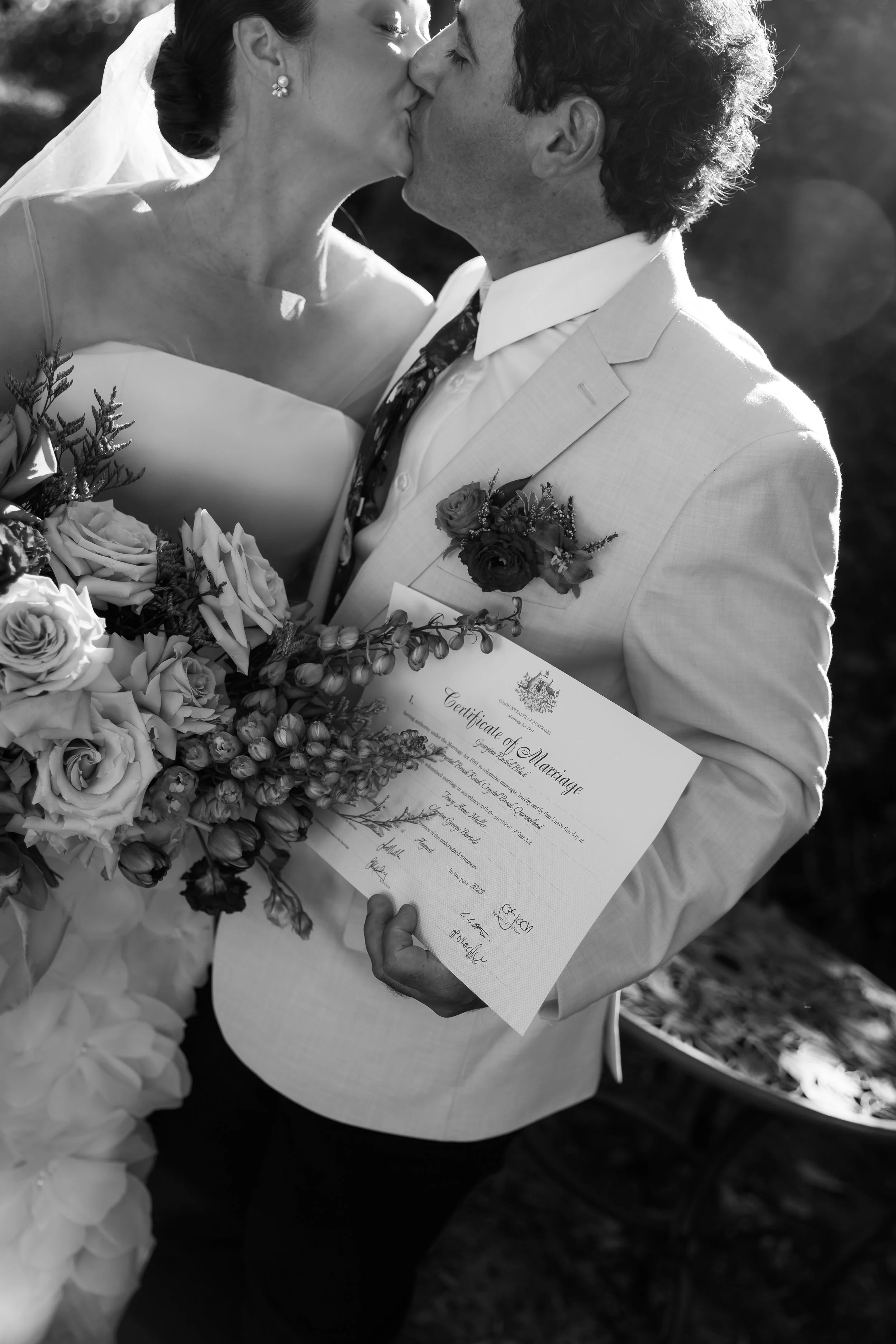 A bride and groom kissing while holding a marriage certificate and surrounded by flowers, outdoors in a wedding setting.