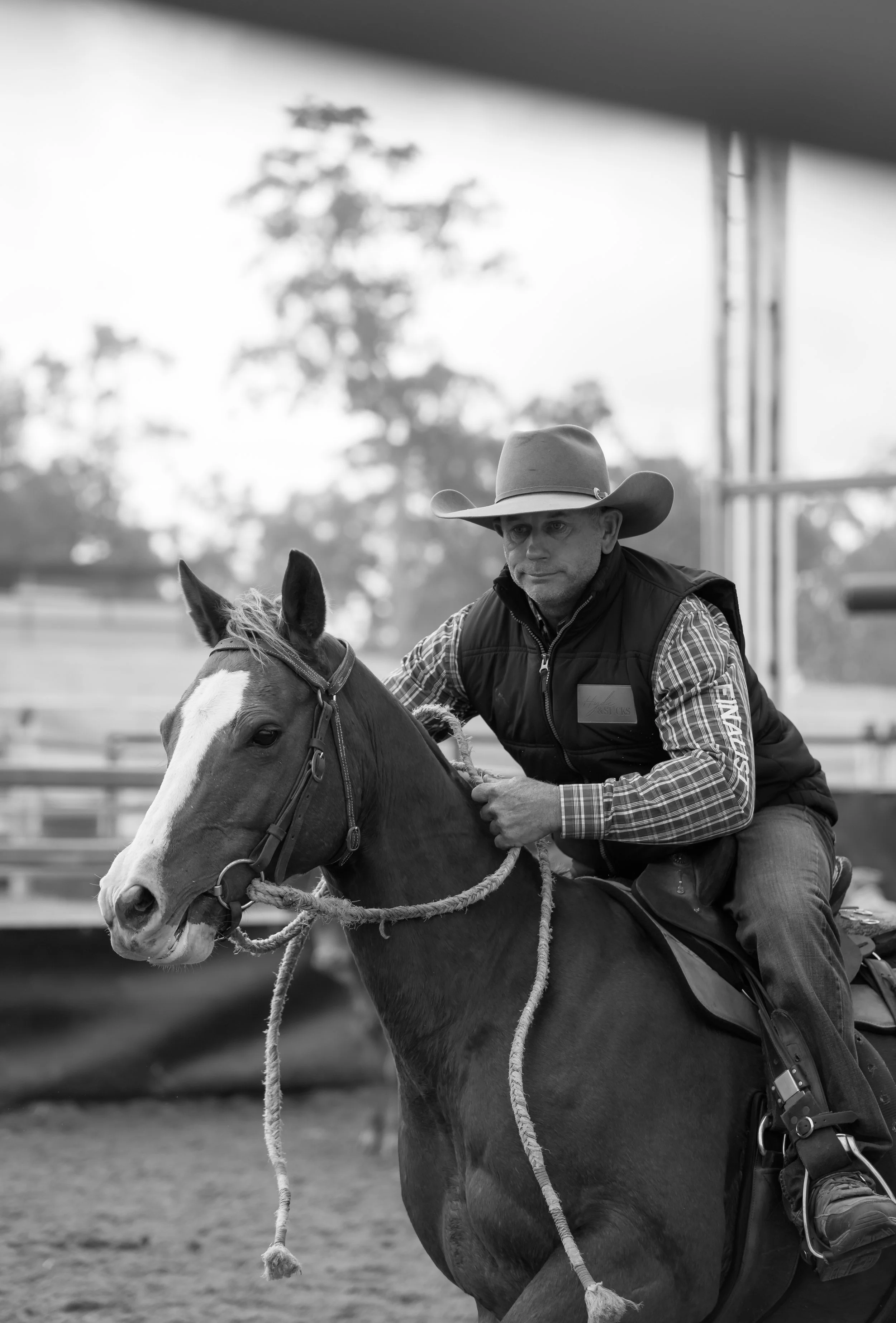 A man wearing a cowboy hat and plaid shirt riding a horse, holding the reins, with a blurred outdoor background.