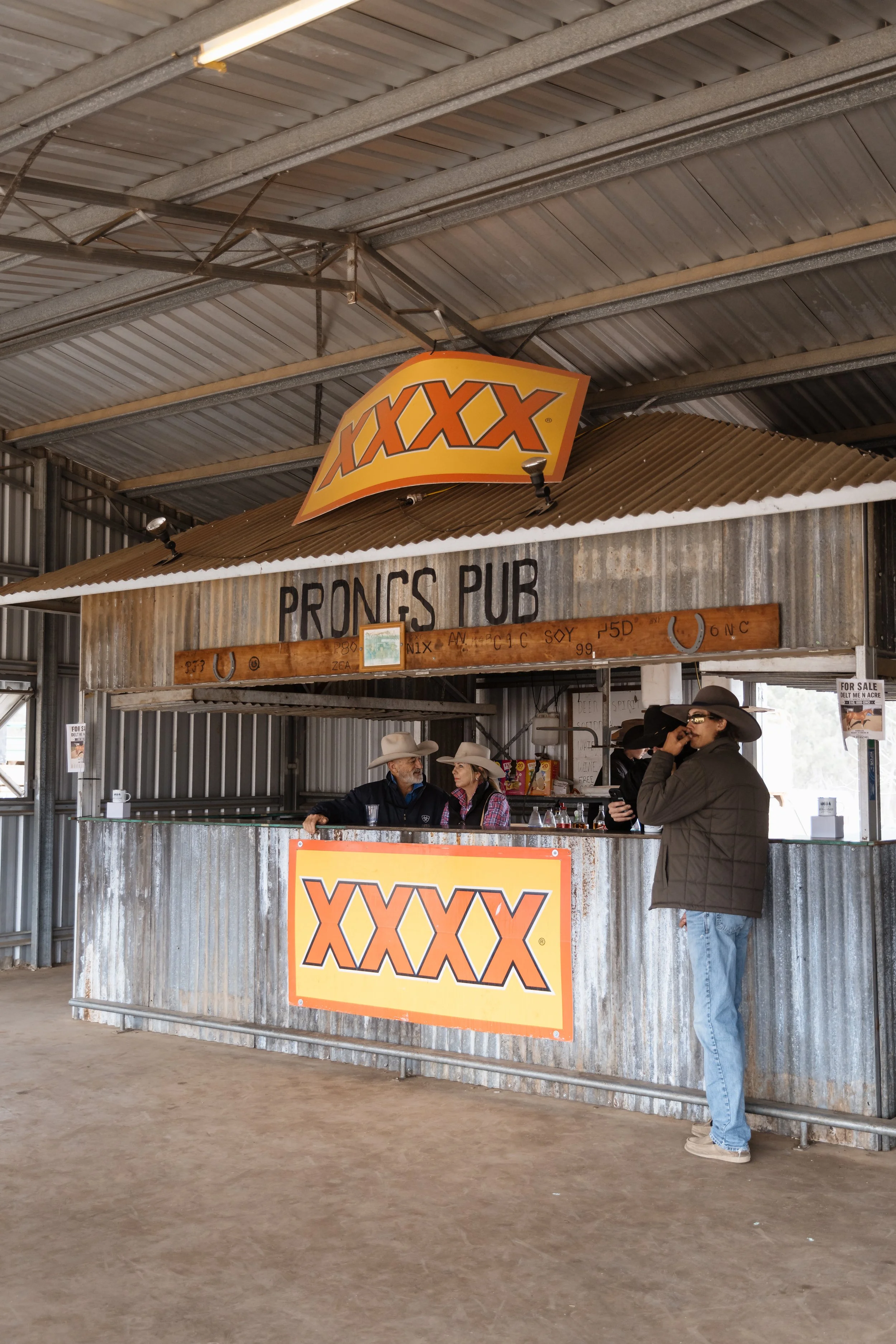 A rustic bar named 'Prongs Pub' with a corrugated metal counter and large 'XXXX' signage. Inside, three people are socializing: two men and a woman, wearing cowboy hats, and a woman in dark clothing wearing sunglasses and talking on the phone.